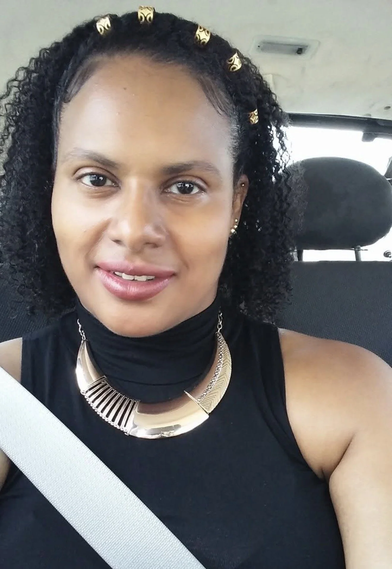 A woman with curly black hair, wearing decorative gold hairpins, a black sleeveless top, and layered gold necklaces, sitting in a vehicle with a seatbelt across her chest.