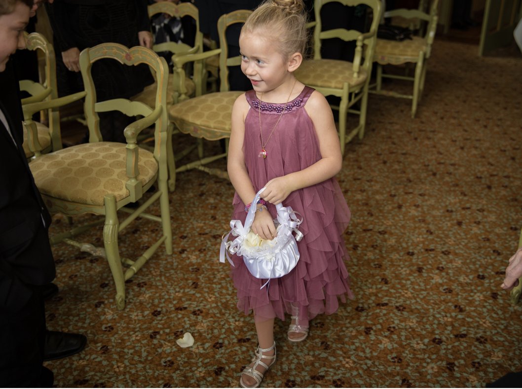 A young girl in a purple dress walking on a patterned carpet, carrying a white basket with ribbons, at a social gathering with chairs.