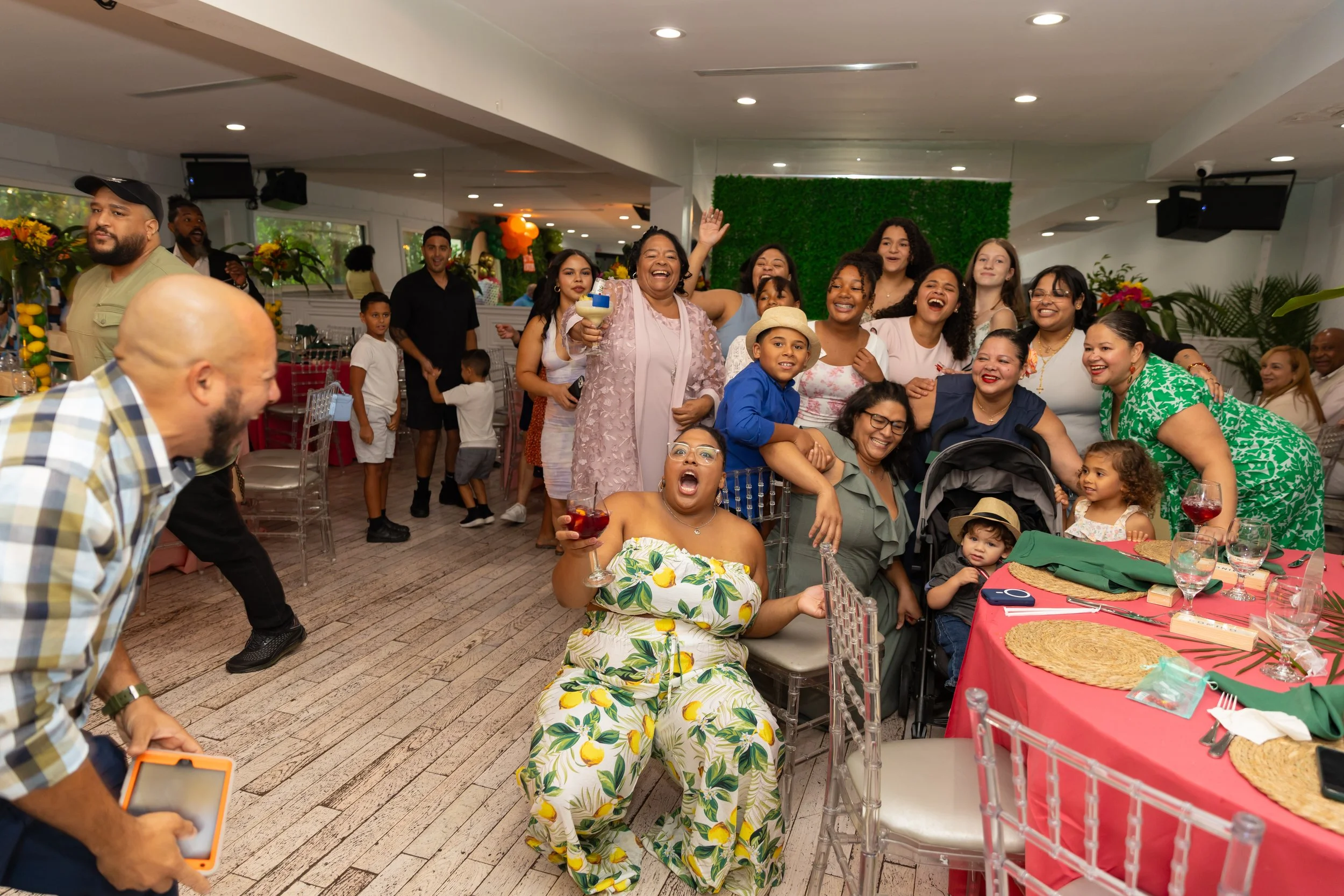 People celebrating at a party, some are smiling and holding drinks, with decorated tables and colorful balloons in the background.