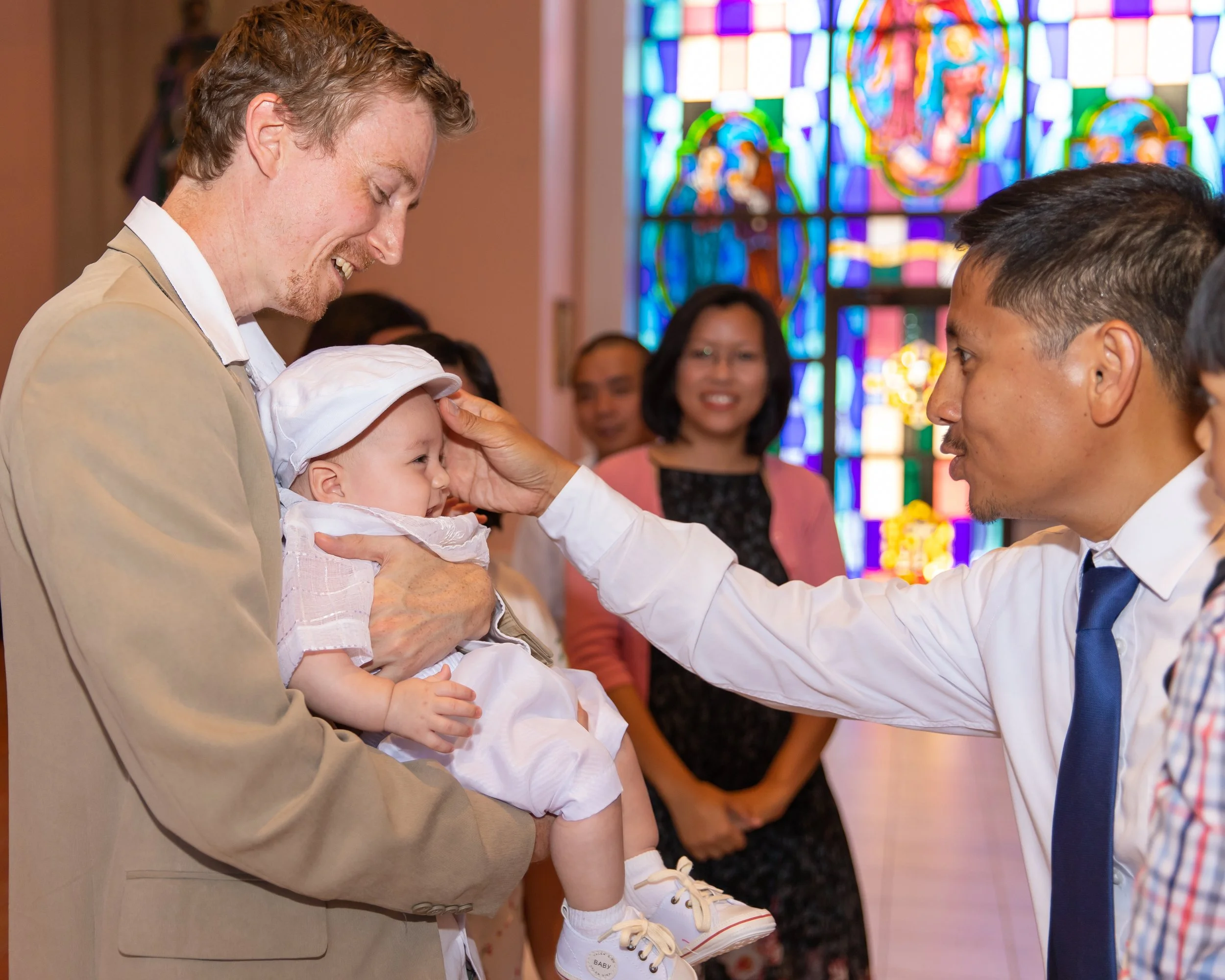 A man in a brown suit holding a smiling baby girl in a white dress and white sneakers during a religious baptism ceremony inside a church with colorful stained glass windows in the background.