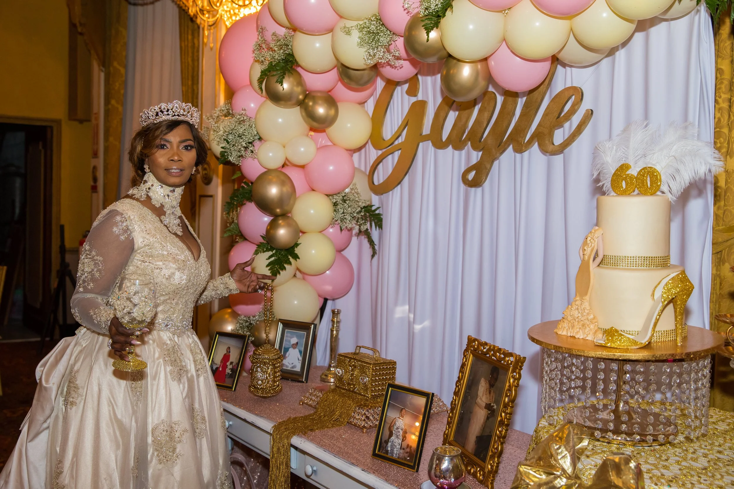 A woman wearing a gold crown and an elegant cream-colored gown stands in front of a decorated table with framed photos and gold accessories. Behind her, there is a balloon arrangement in pink, cream, and gold, alongside a cake decorated with gold acc