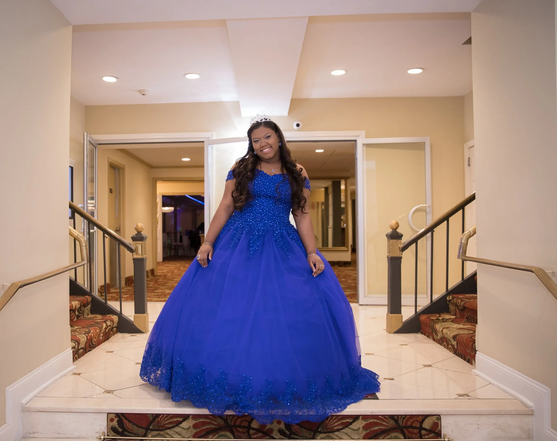 A young woman in a royal blue ball gown with lace details and a tiara standing on a staircase in an elegant hotel lobby.