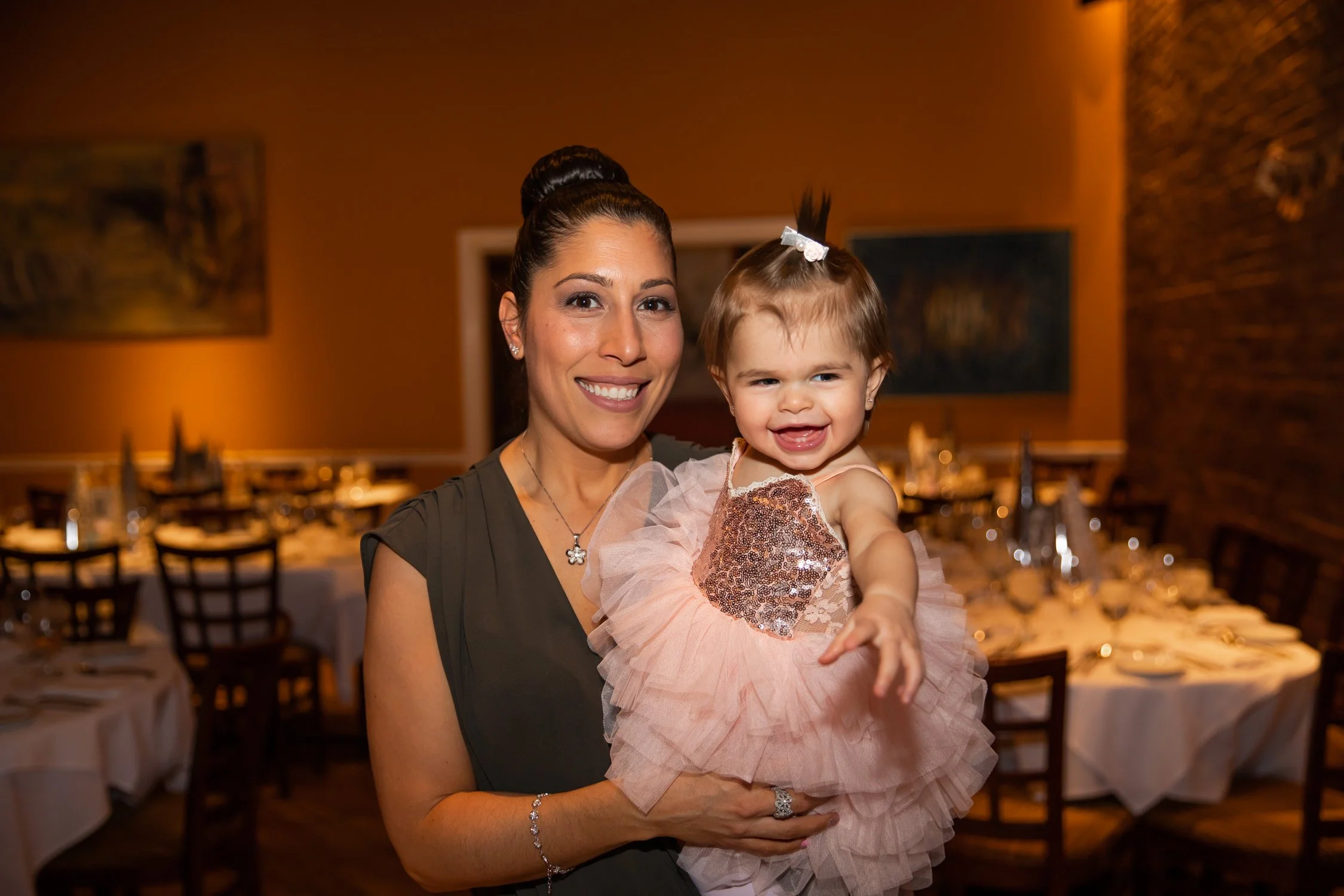 A woman smiling and holding a young girl dressed in a pink, sequined dress in a warmly lit restaurant or banquet hall with tables and chairs set for an event.