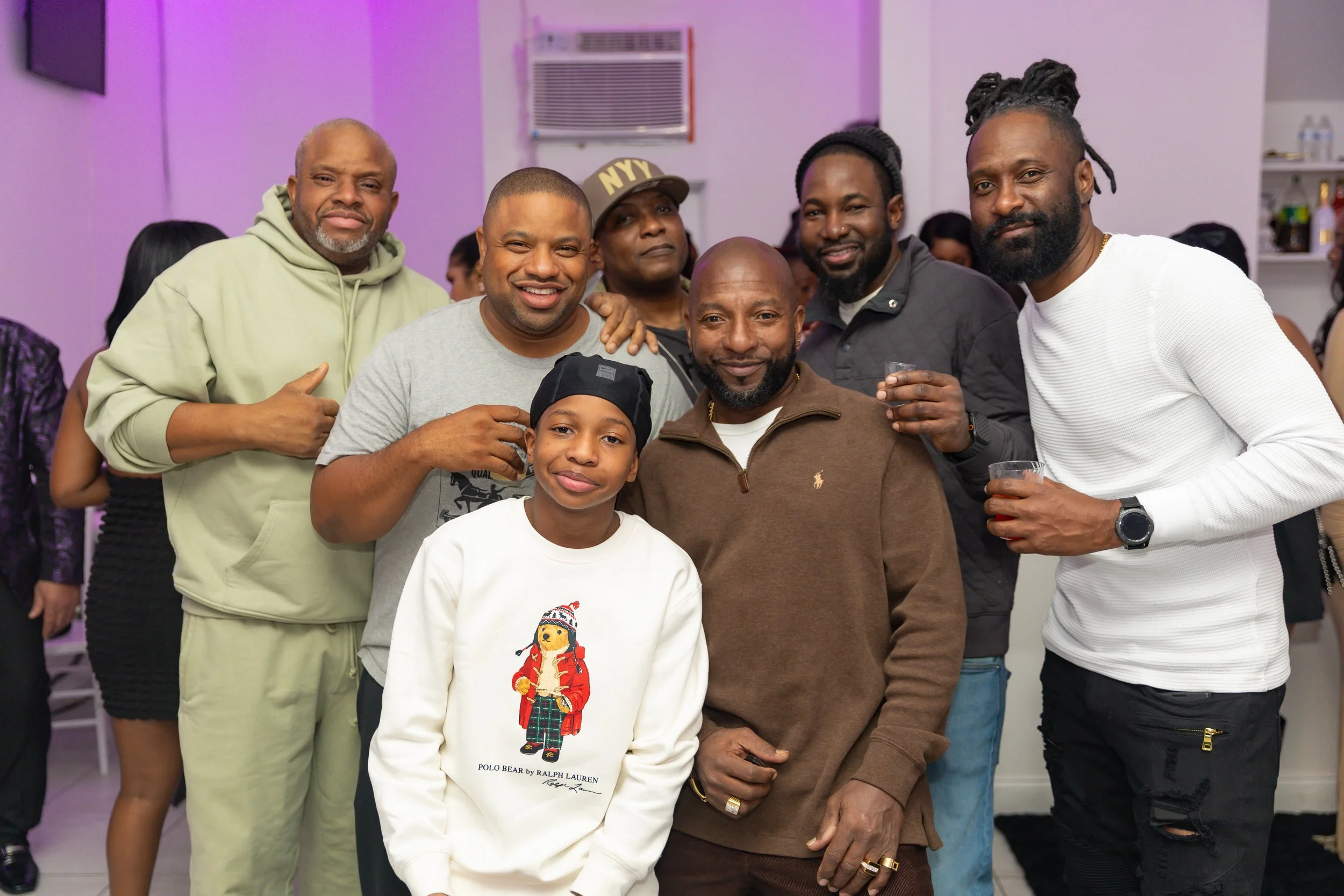 A group of seven smiling people, including a child, gathered in a room with purple lighting, posing for a photo. They appear happy and friendly.