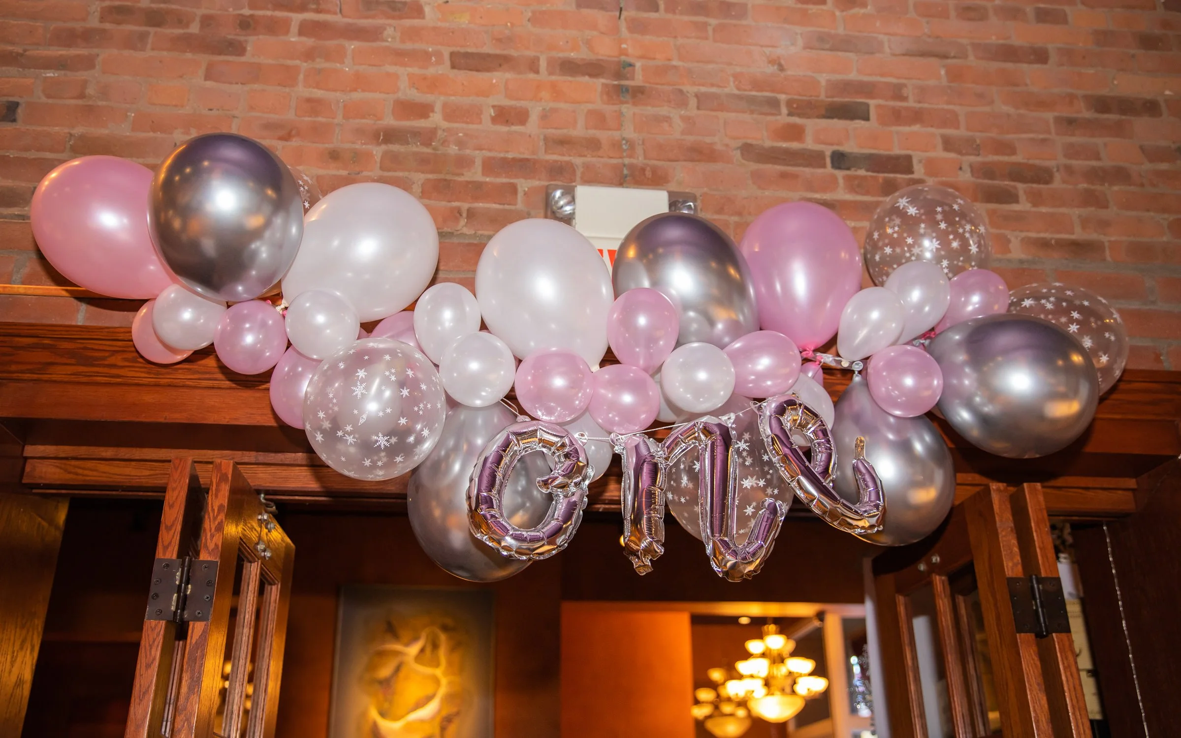 A balloon decoration with pink, white, silver, and transparent balloons with white snowflake patterns, forming the word 'LOVE' in shiny metallic balloons, against a brick wall backdrop.