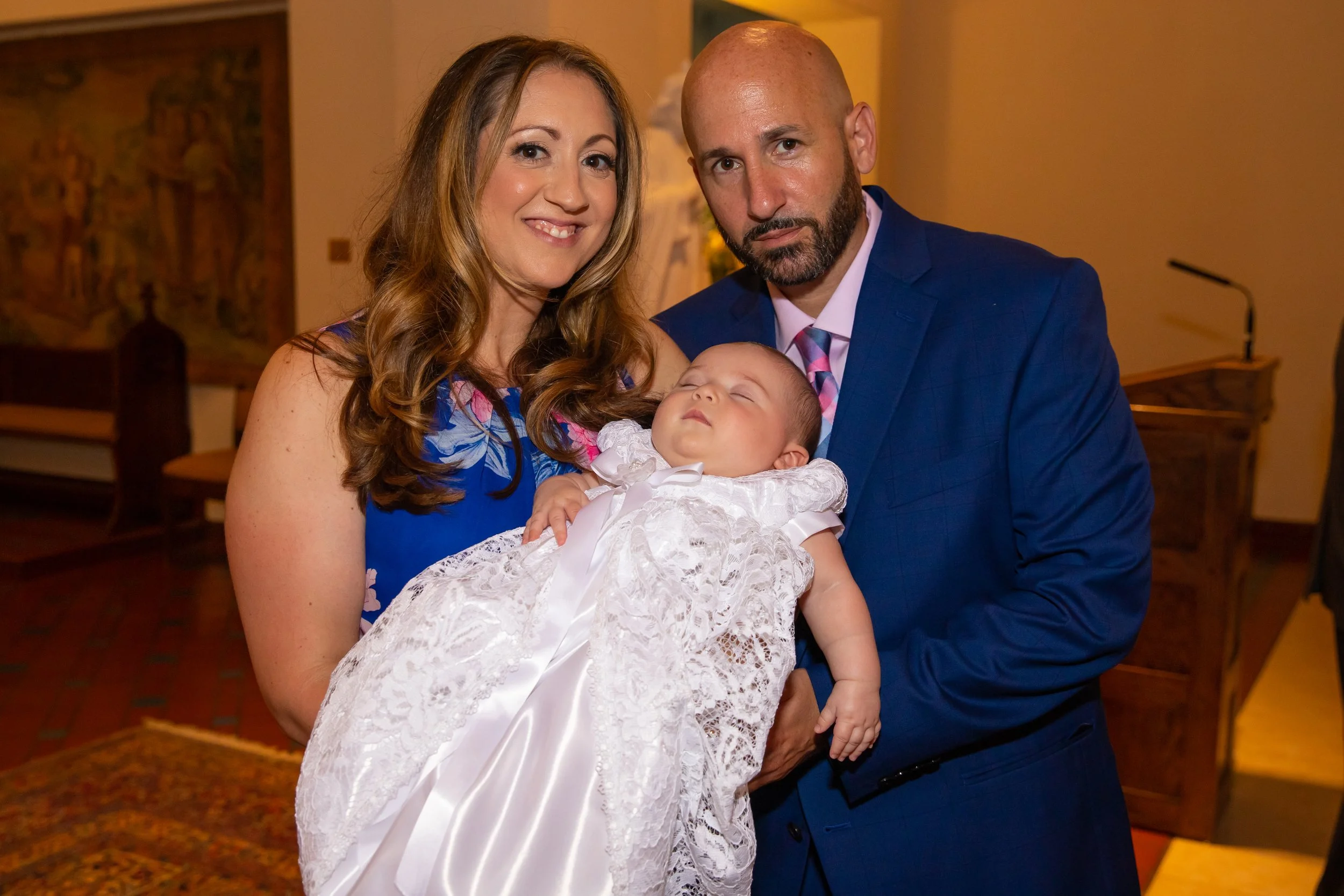 A woman in a blue dress, a man in a blue suit, and a baby in white dress are posing for a photo indoors.