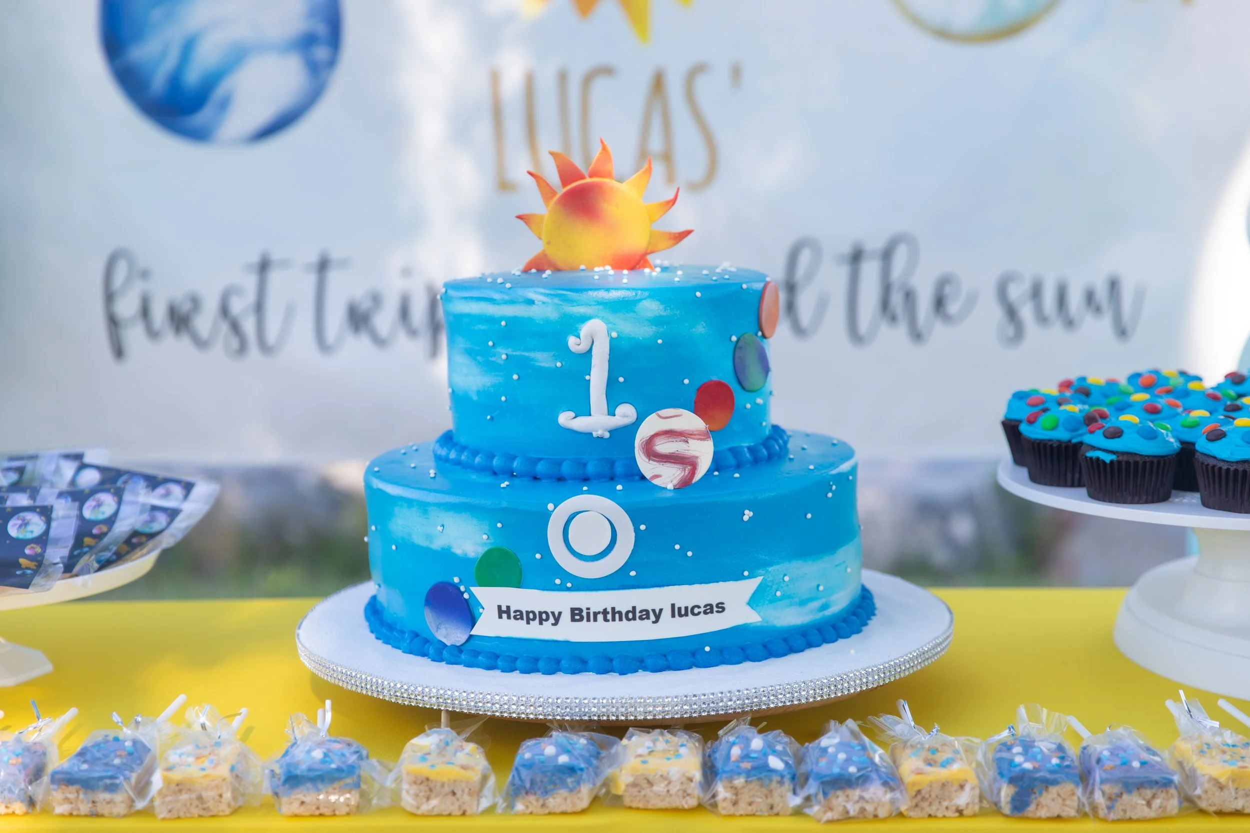 A blue birthday cake decorated with colorful circles, white sprinkles, and a flaming orange and yellow fondant sun on top, with a sign that reads "Happy Birthday Lucas." Surrounding the cake are small, square candies with blue and white sprinkles, an