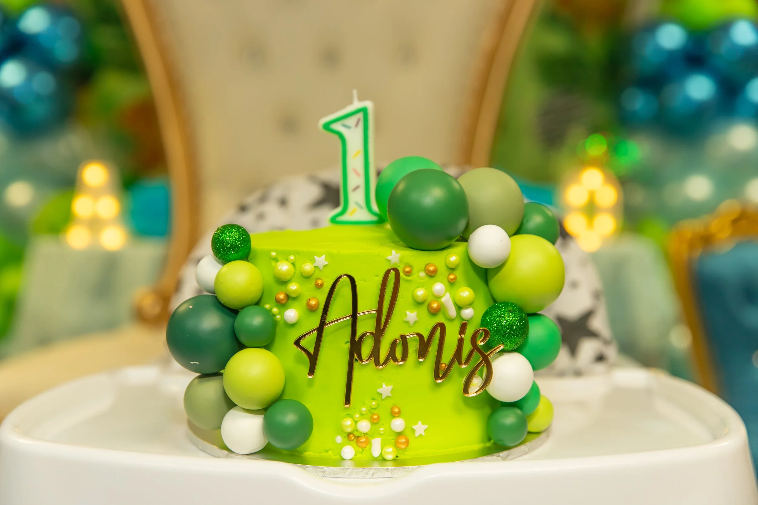 A green birthday cake decorated with white, green, and gold balloons and a large illuminated number one candle, with a mirror back and blurred fairy lights and decorations in the background.