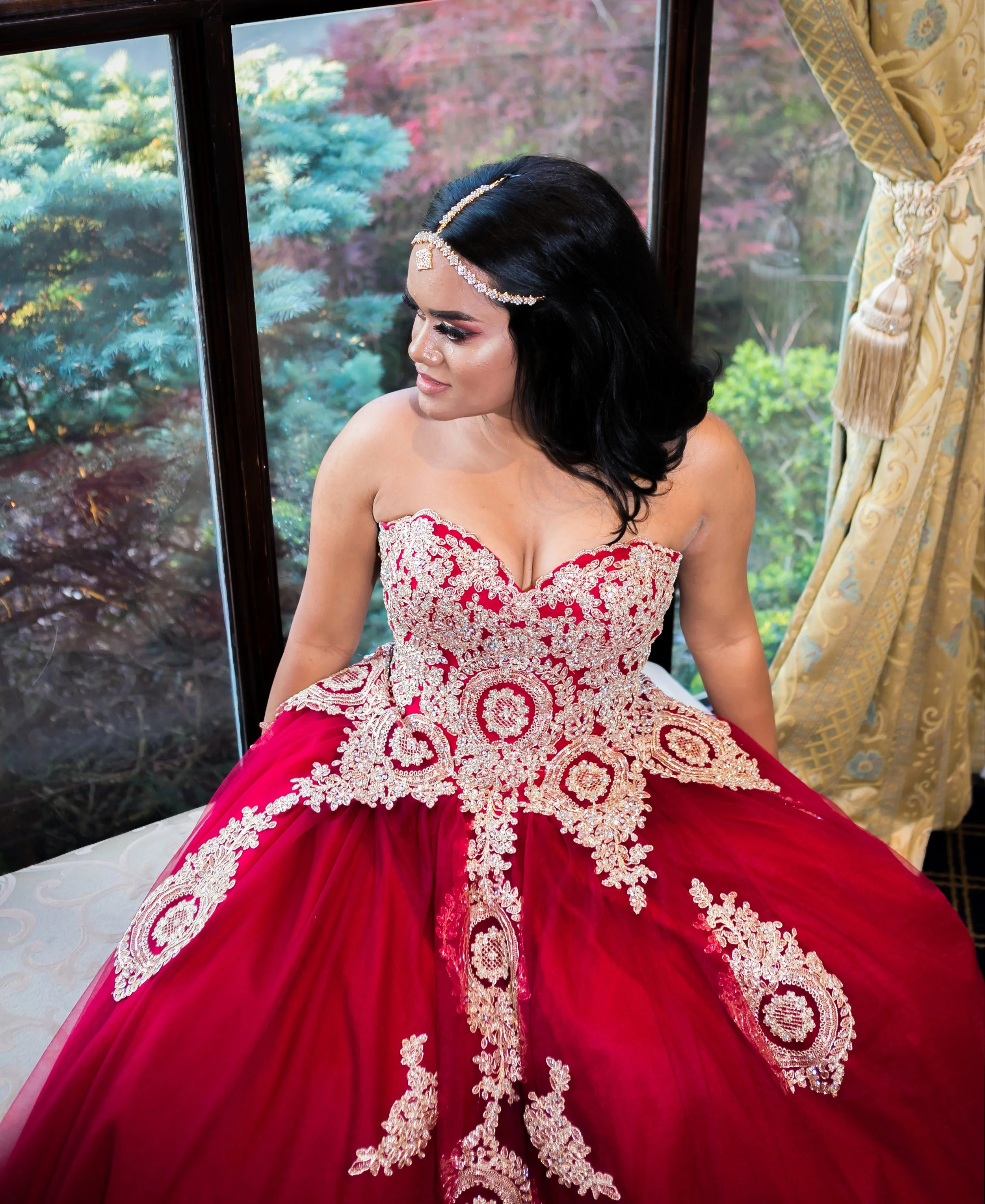 A woman with black hair wearing an elaborate red dress with gold embroidery and holding the skirt out, sitting near a window with a view of trees outside.