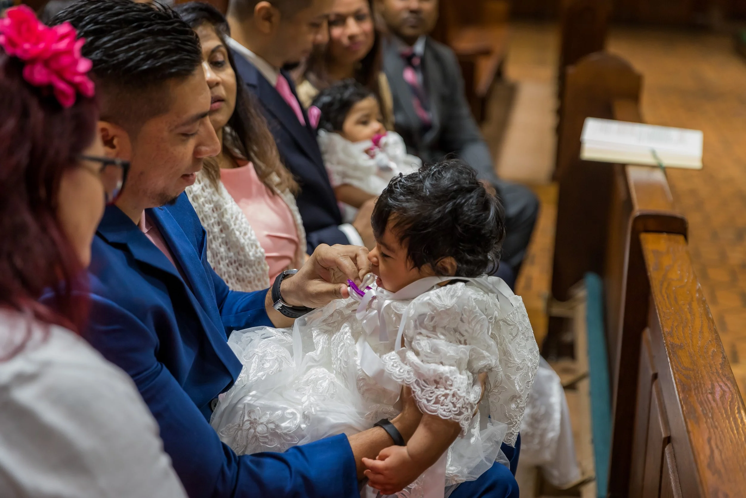 A young girl dressed in a white lace dress with a ribbon in her hair, sitting on a man's lap, receiving a blessing or prayer from a man in a blue suit. They are seated on wooden church pews, with other similarly dressed people sitting behind them, in