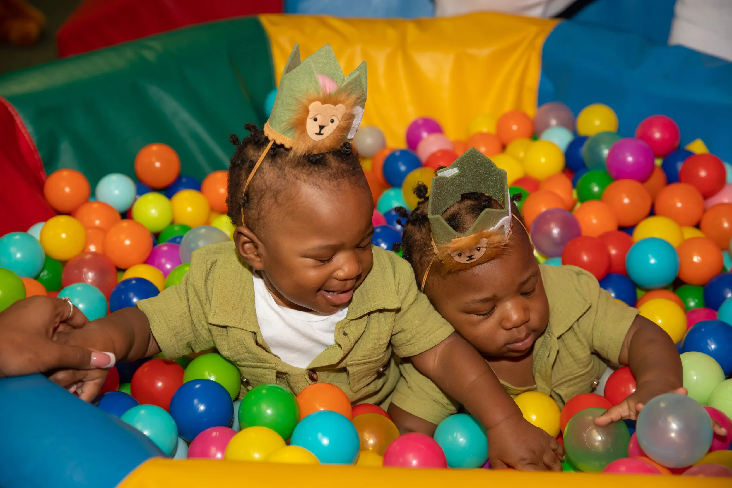 Two children playing in a colorful ball pit, wearing headbands with lion faces on them.