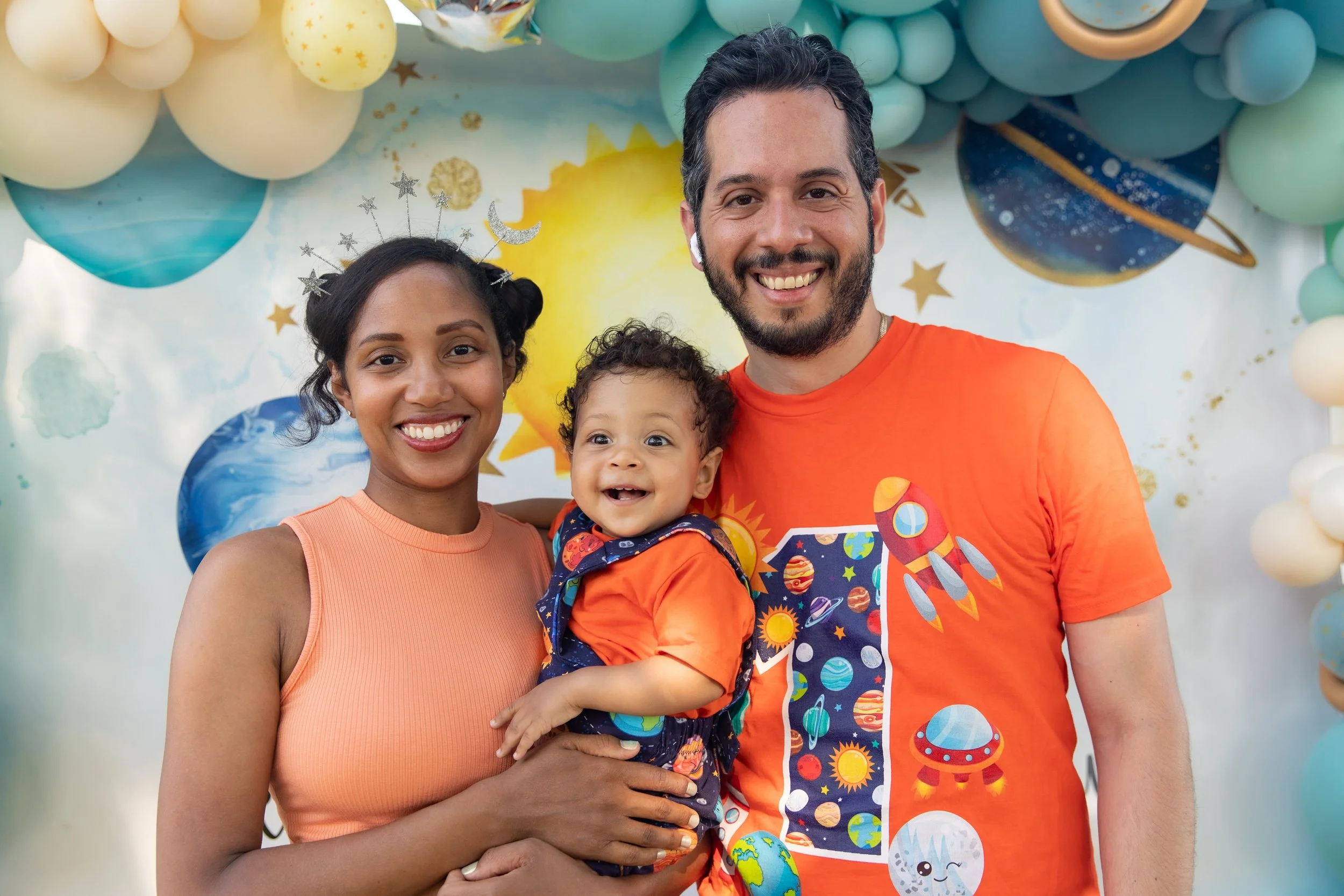 A happy family of three standing close together, smiling, against a colorful space-themed backdrop with balloons and planets. The mother has dark hair in buns and wears a peach top, the father has dark hair and beard, wearing an orange shirt with spa