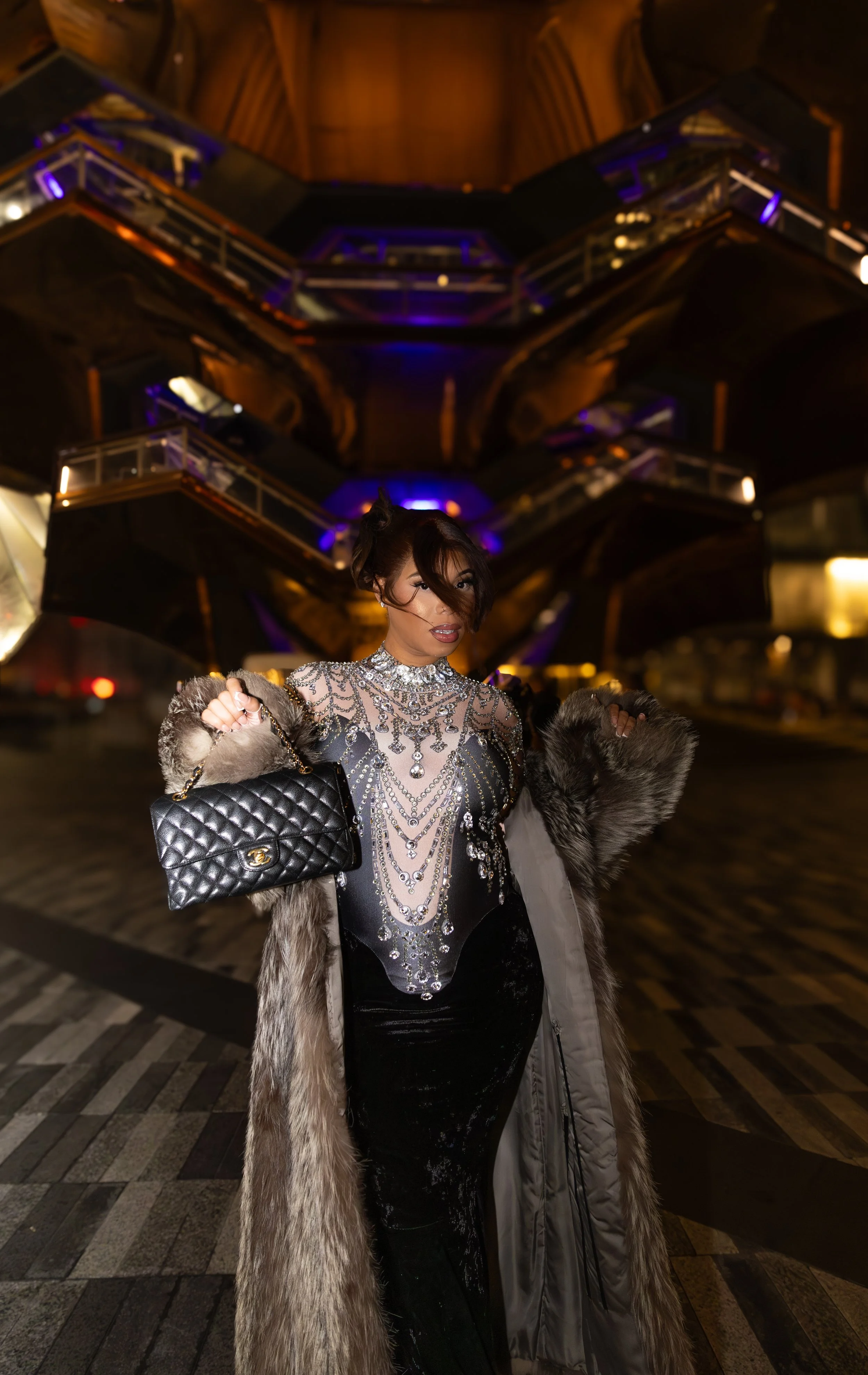 Woman in glamorous, jeweled top, wearing a fur coat, carrying a quilted black purse, standing in front of modern, illuminated building at night.