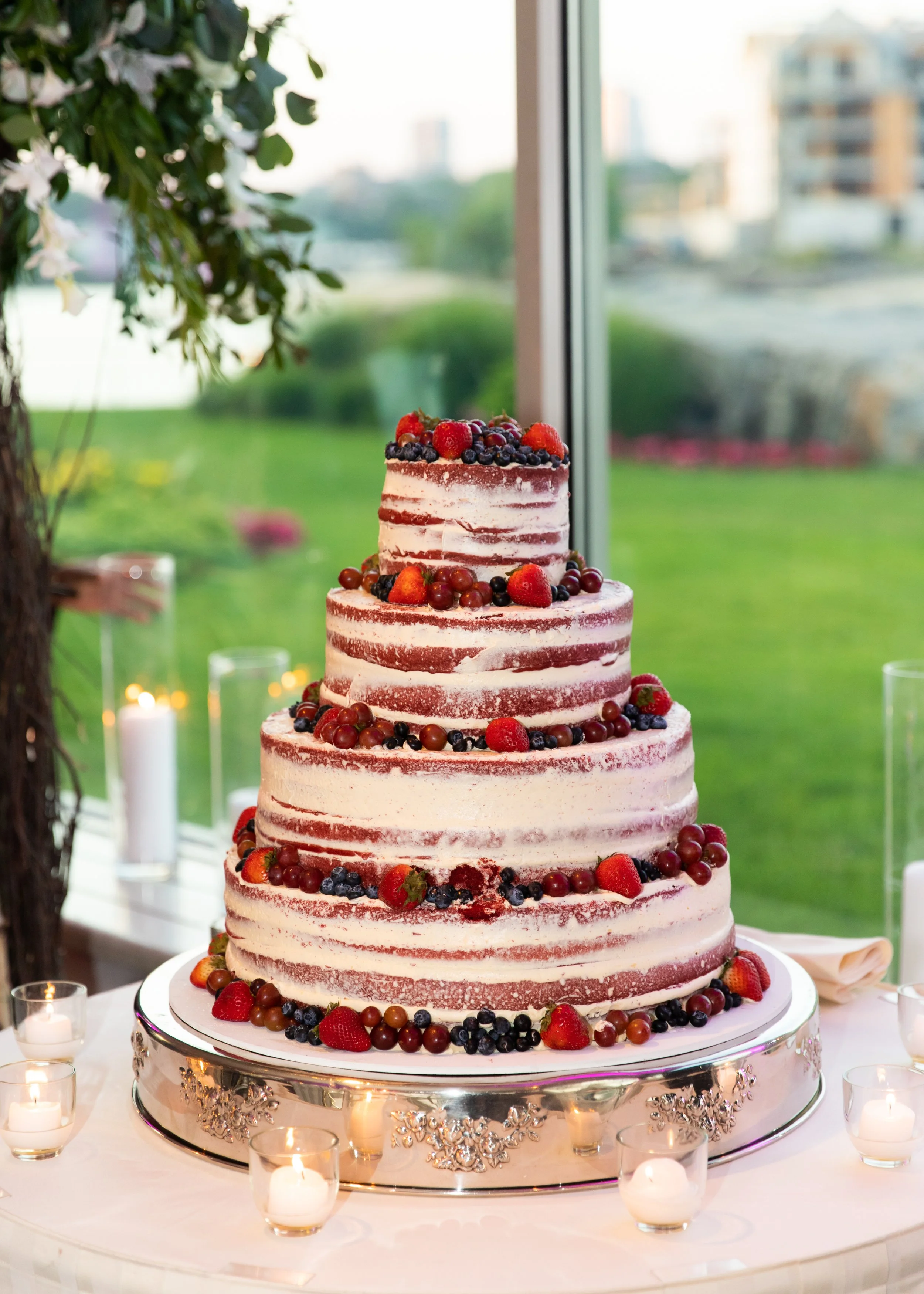 A large, multi-layered wedding cake with white frosting and red velvet layers, decorated with fresh strawberries, blueberries, and grapes on top and around the edges, placed on an ornate silver cake stand surrounded by small lit candles.