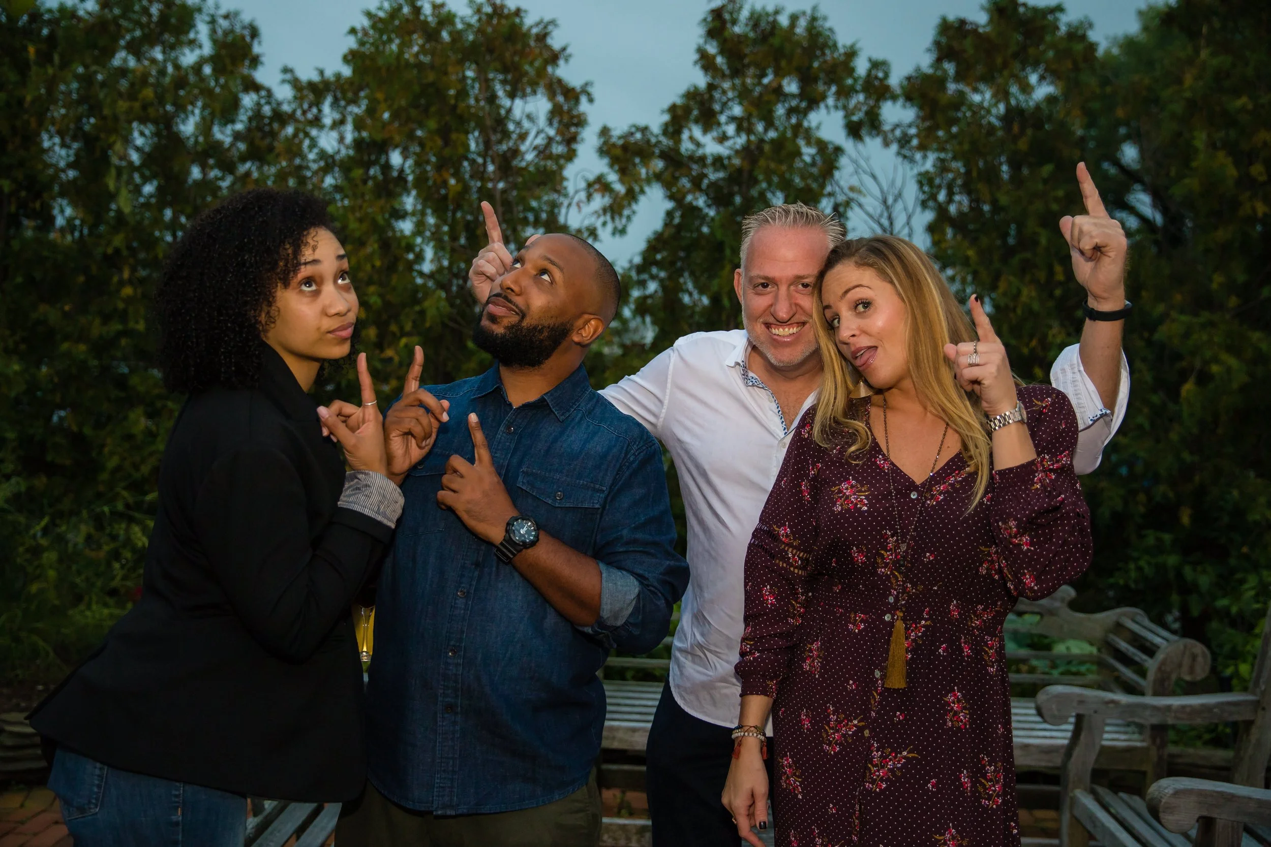 Four people, two men and two women, standing outdoors with trees in the background. They appear cheerful, with three of them pointing upwards and one with her tongue out.
