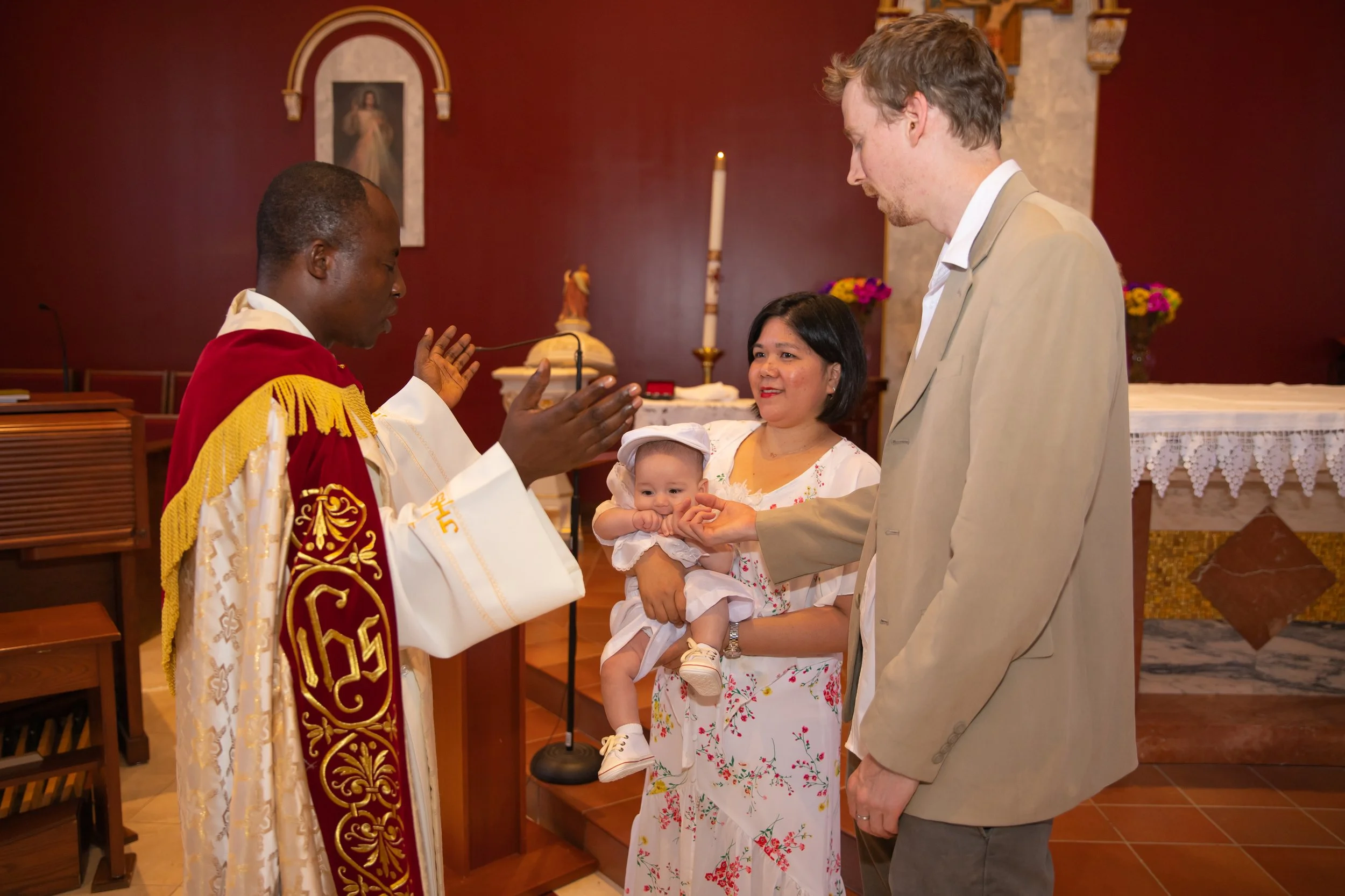A priest dressed in ornate robes performs a baptism ceremony for a baby held by a woman, with a man standing beside her, inside a church with religious artwork and flowers in the background.