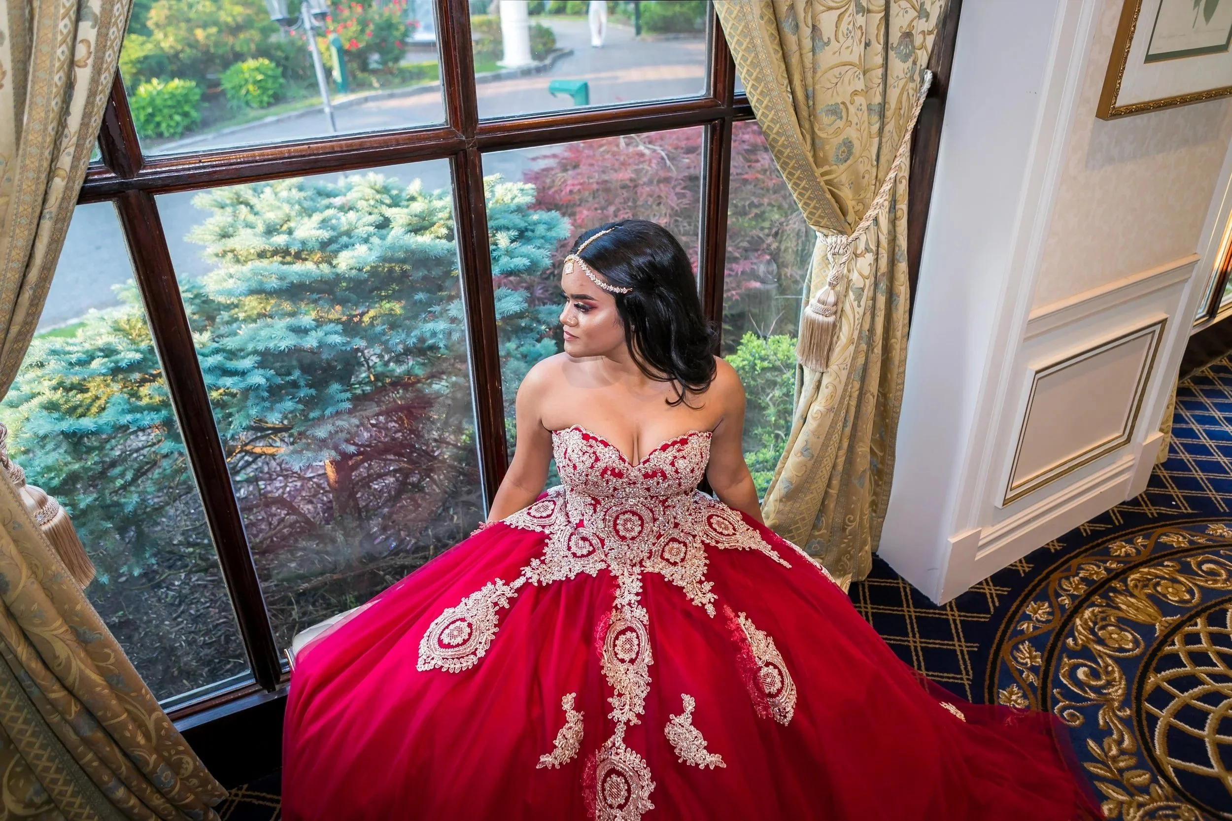 A woman in a red and white embroidered dress with a strapless design sits by a large window with gold curtains, looking to the side. Outside, there are lush green trees and bushes.