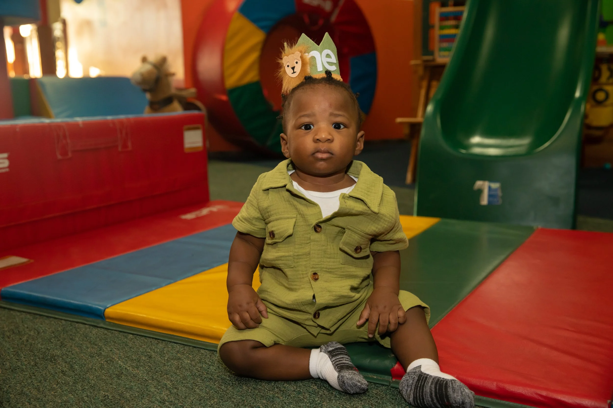 A young boy sitting on the floor at an indoor playground, with colorful padded play structures and a plush lion toy in the background.