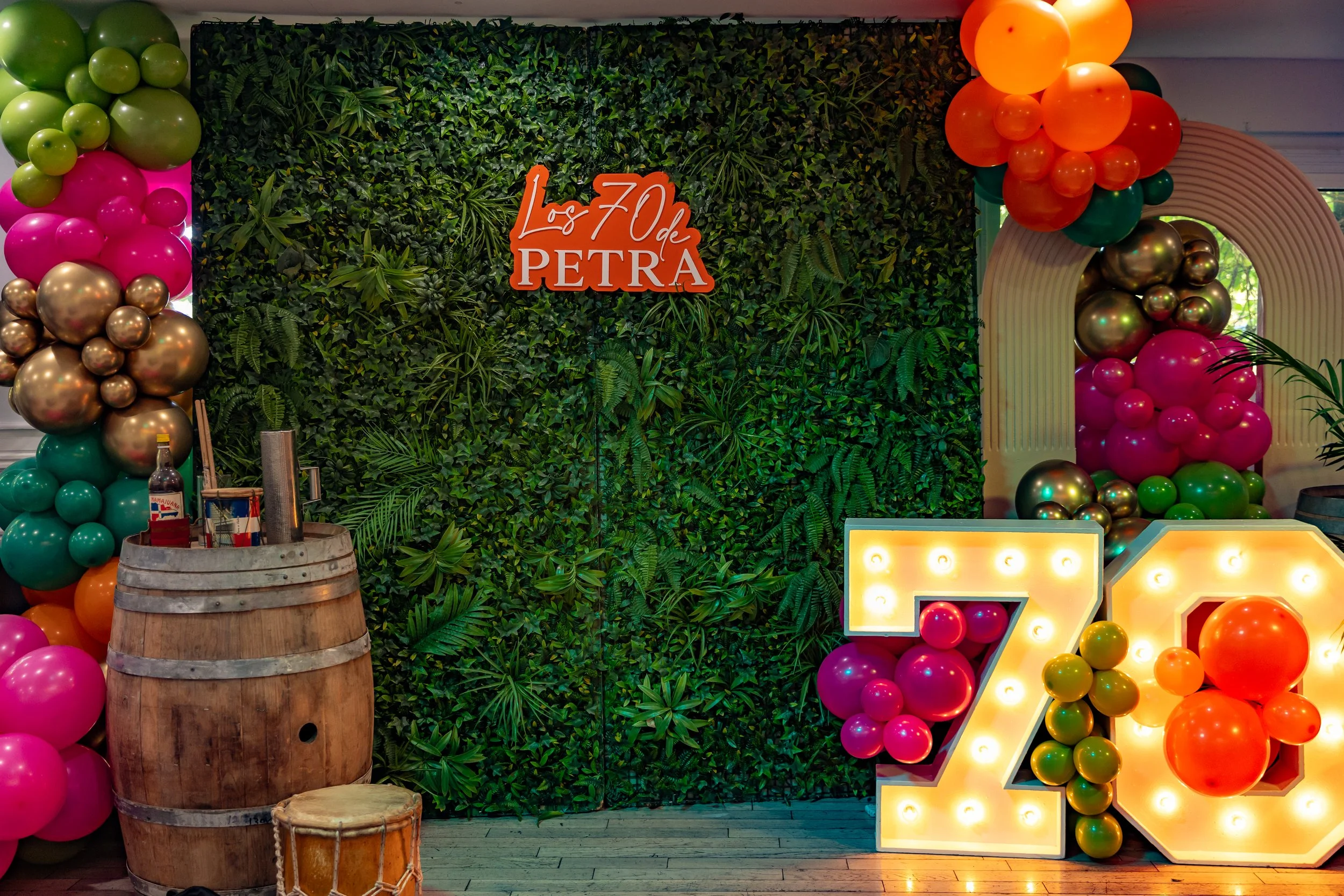Decorative backdrop with green foliage, pink, gold, orange, yellow, and green balloons, a large illuminated number 7, a barrel with a bottle of liquor, and a drum in front.