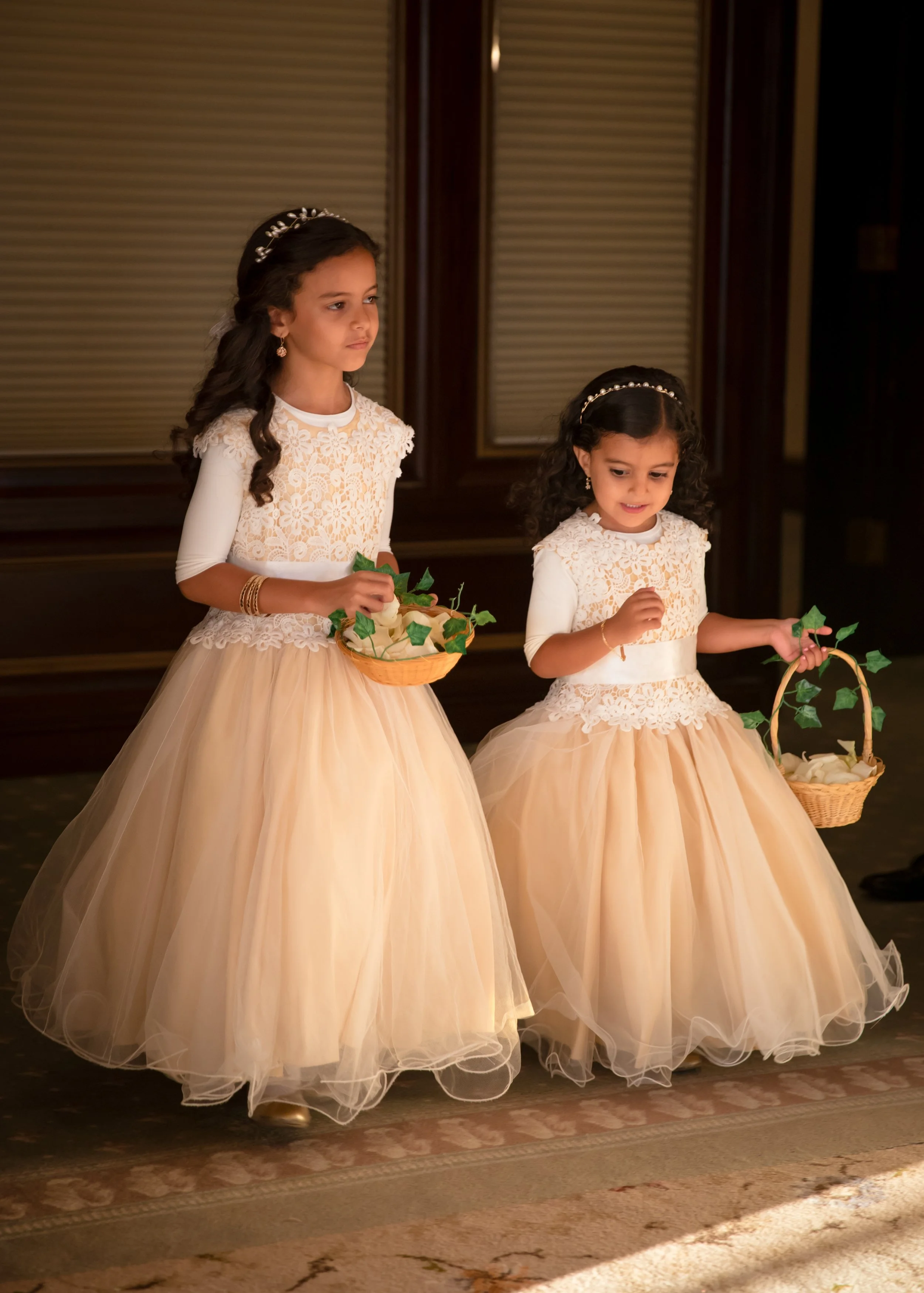Two young girls in cream-colored lace and tulle dresses, each wearing a pearl headband and earrings, standing indoors holding small baskets with artificial flowers.