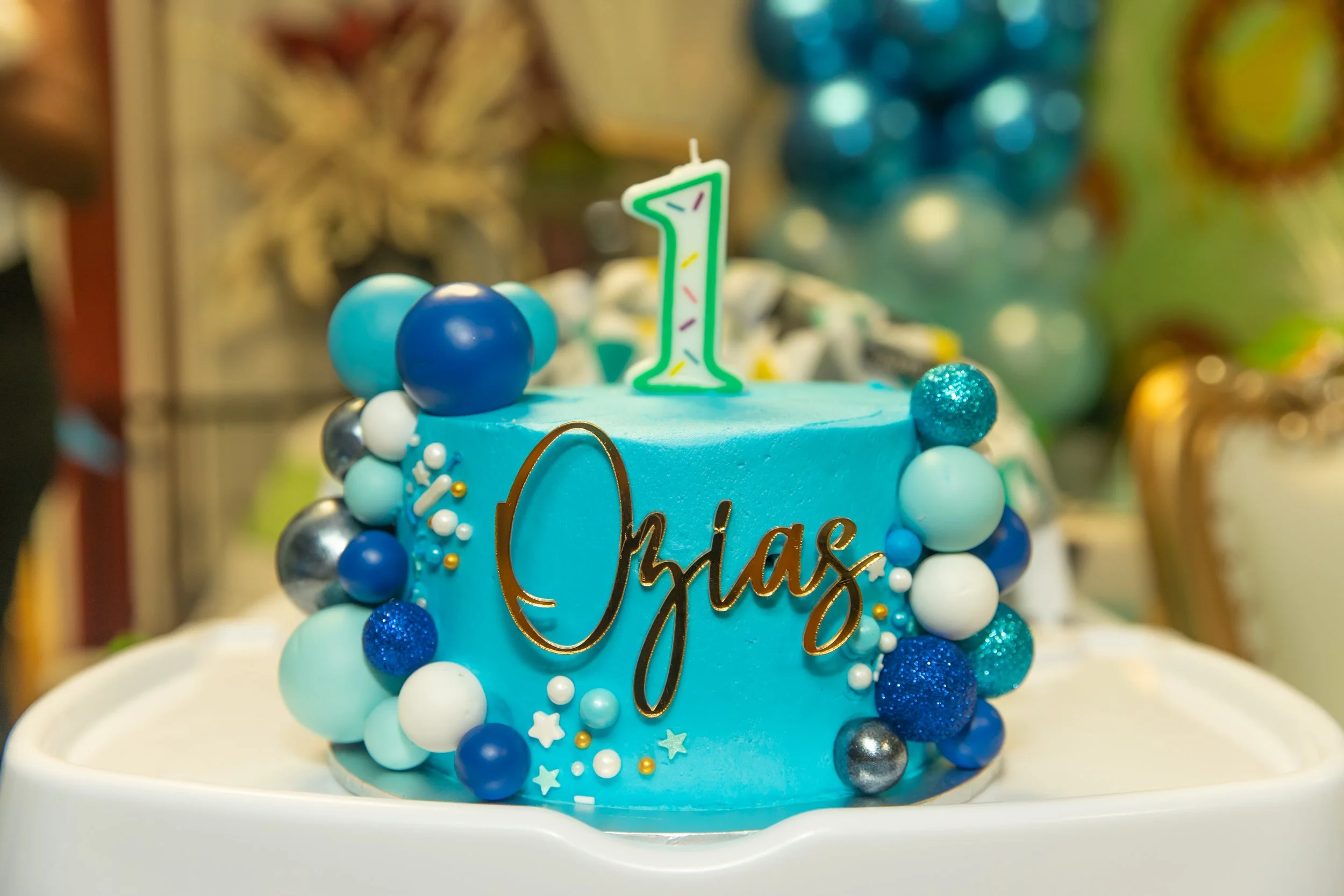 A blue birthday cake decorated with blue and white spherical ornaments, a glittery blue ball, and a green candle shaped like the number 1. The cake has gold writing and star-shaped decorations. The background features blurred colorful decorations.