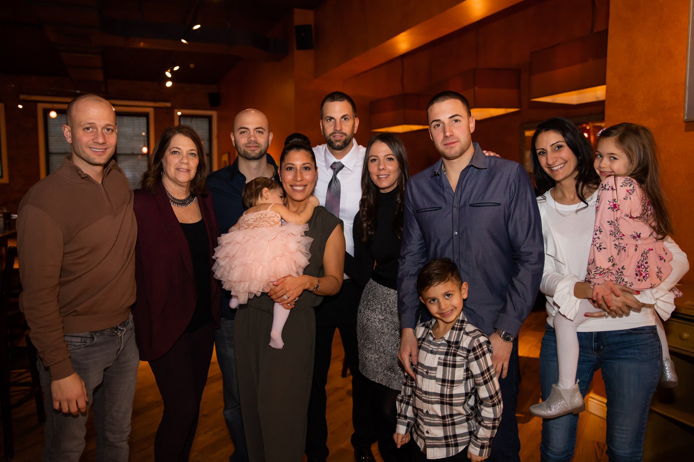 A group of eleven people, including adults and children, pose together in a warmly lit indoor setting. They are smiling and standing close to each other.