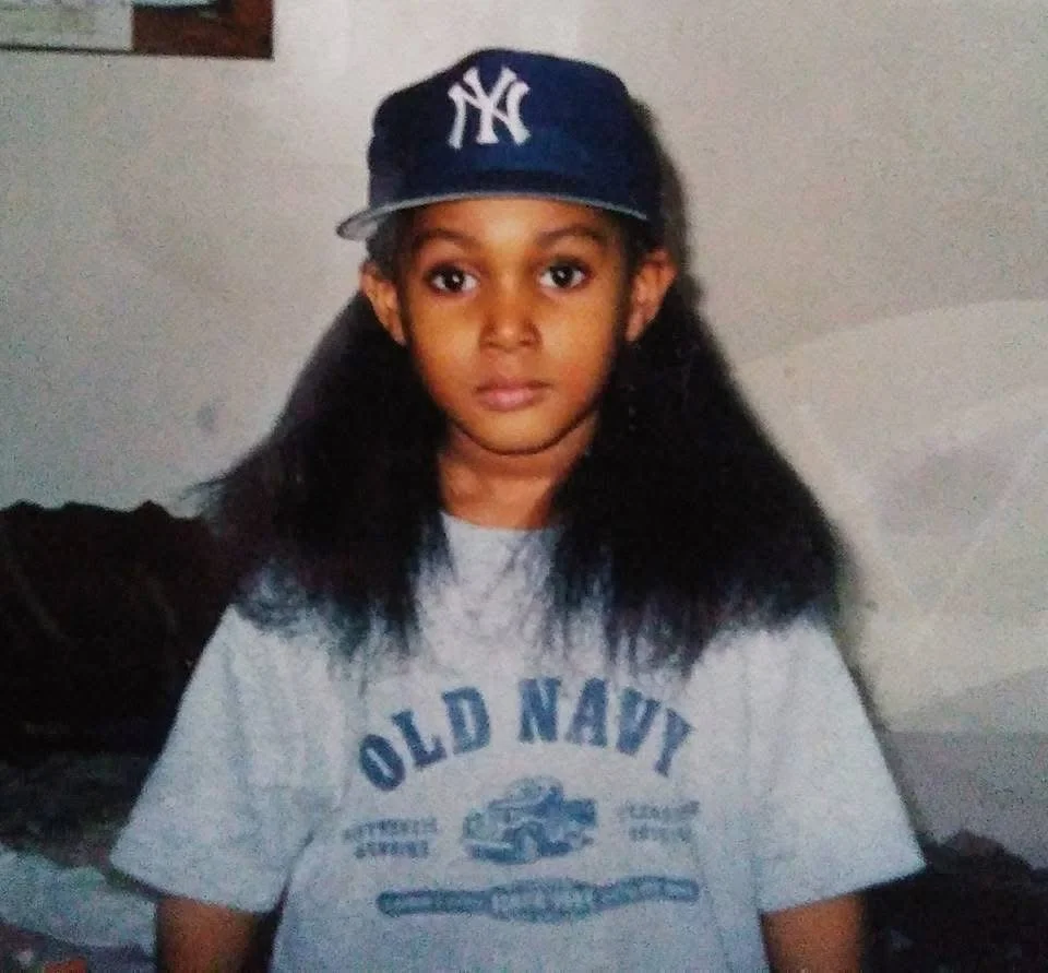 A young girl with long black hair wearing a navy blue New York Yankees cap and a gray Old Navy t-shirt, standing indoors with a neutral expression.