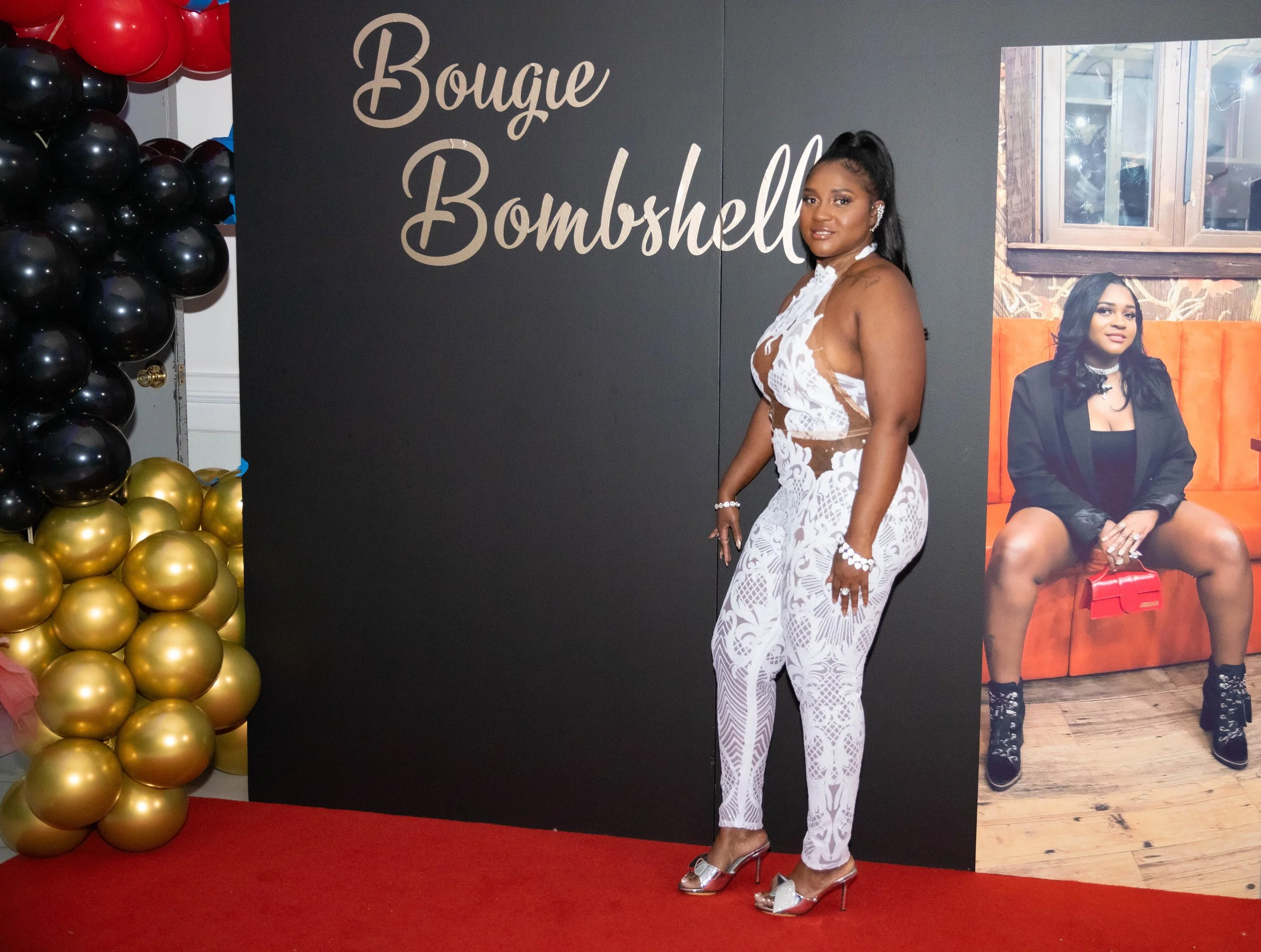 A woman in a white lace outfit with high heels stands on a red carpet at an event, with a backdrop that has the words "Bougie Bombshell" and black and gold balloons on the side.