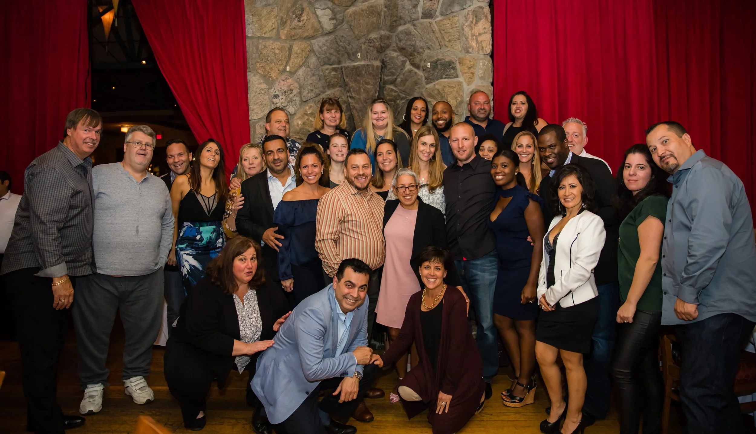 A large group of diverse people gathered together, smiling, in a setting with a stone wall and red curtains in the background.