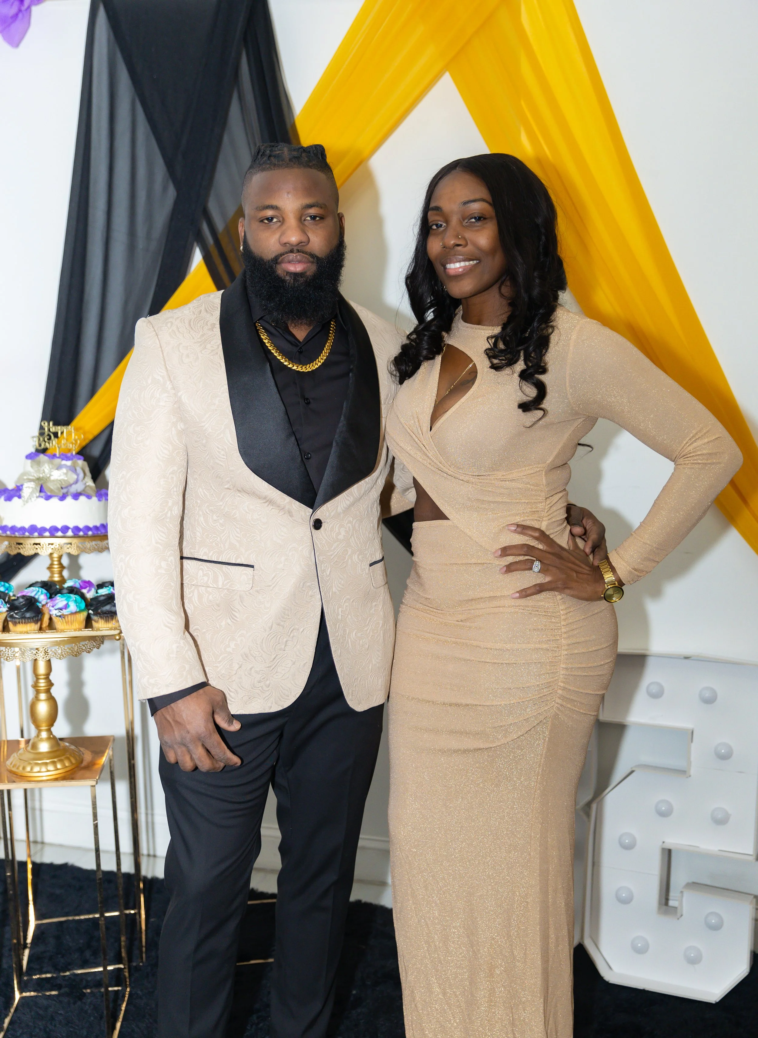 A man and a woman dressed in formal attire standing close together at a celebration, with a decorated cake and party decorations in the background.