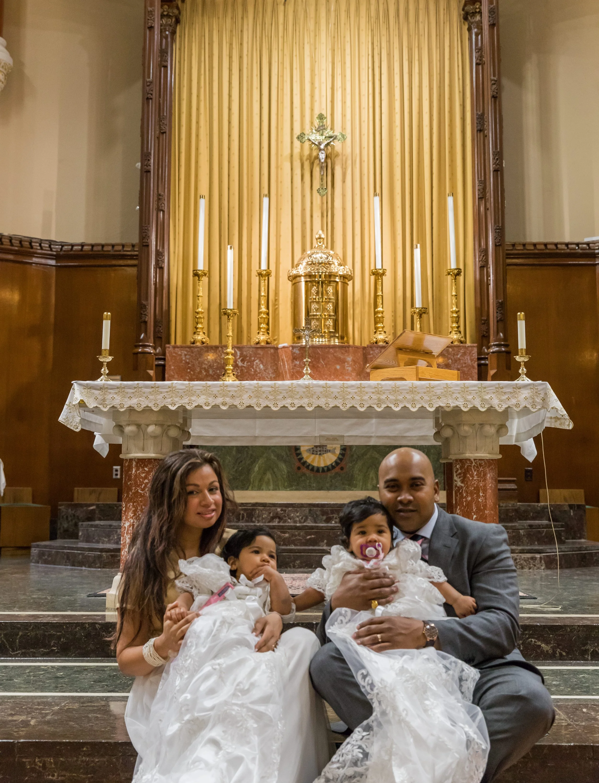 A family dressed in formal attire sitting on steps in front of an altar with a crucifix, candles, and gold decorations inside a church.