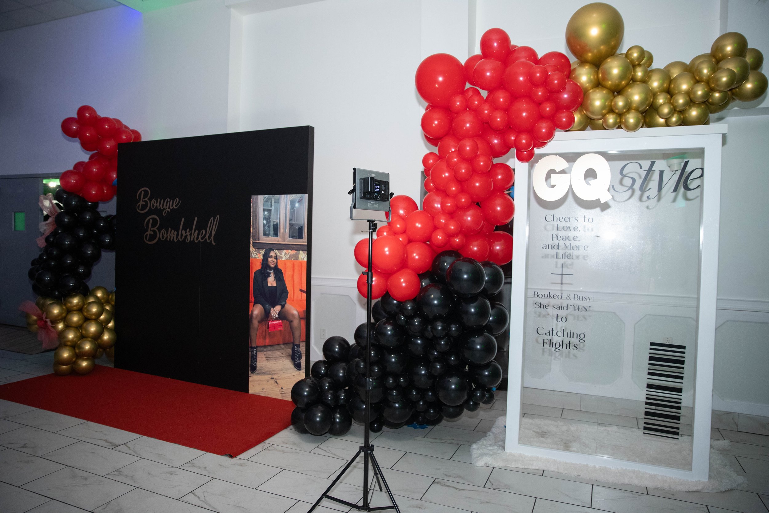 Decorative setup with red, black, and gold balloons forming arches and clusters at an event. There is a red carpet, a standing photo booth with a woman seated on a red couch, and signs with text, including 'GQ Style' and 'Bougie Bombshell.'