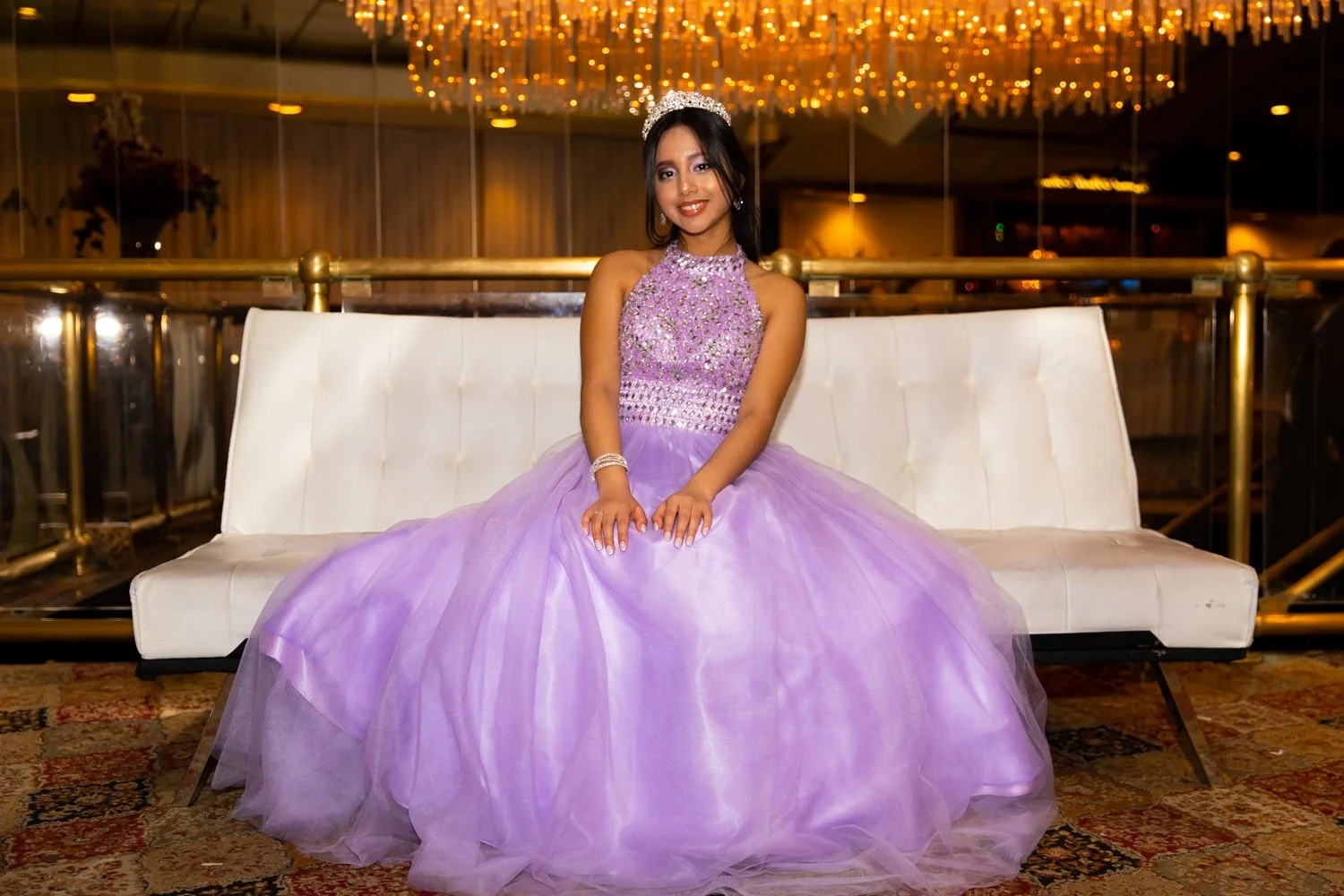 A young woman in a lavender ball gown with a bejeweled bodice and a sparkly tiara, sitting on a white bench in a fancy indoor setting with warm lighting and a chandelier above.