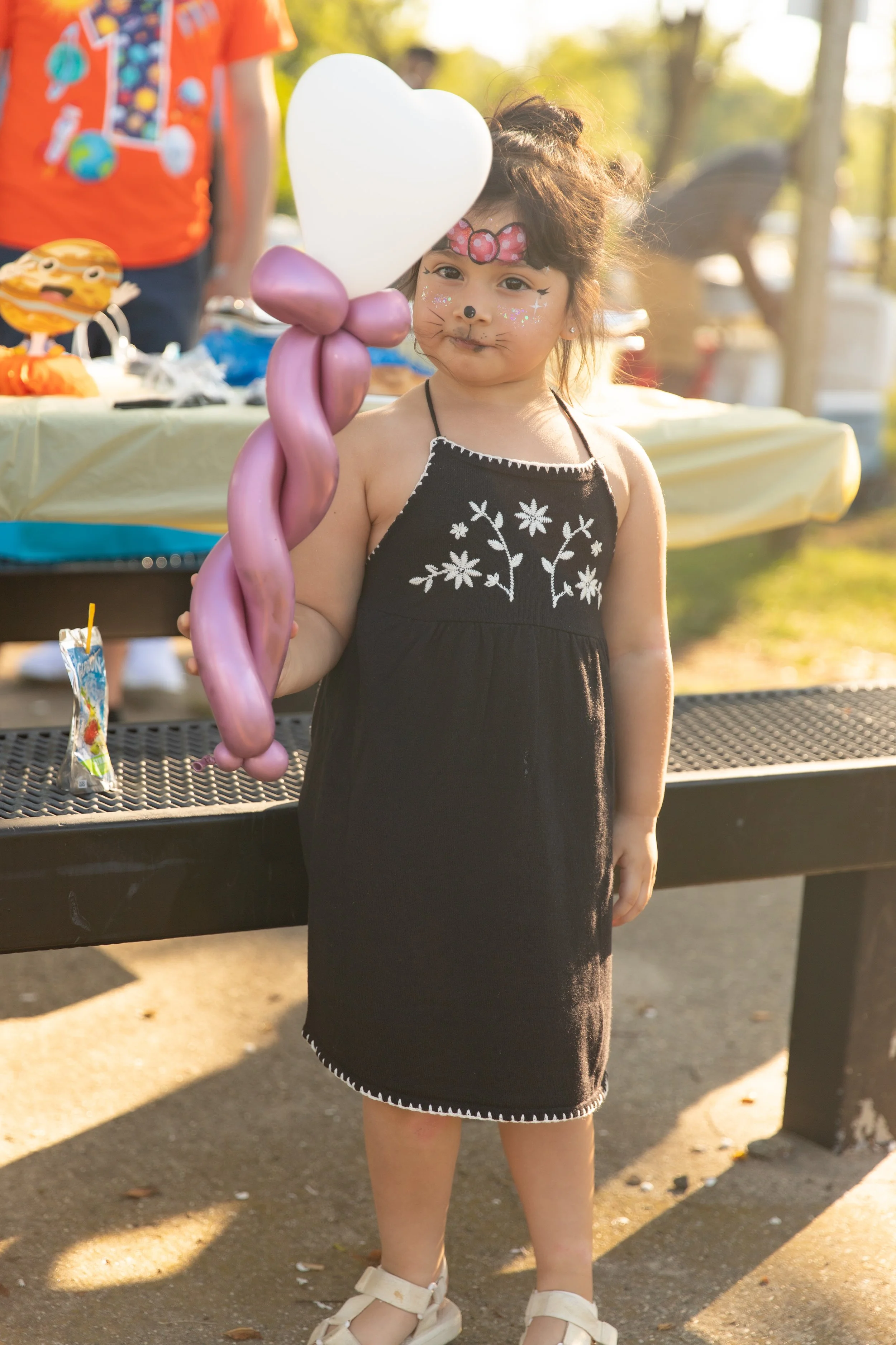 Young girl with face paint and a pink bow headband holding a cluster of balloons at an outdoor event, with a table of items in the background and sunlight filtering through trees.