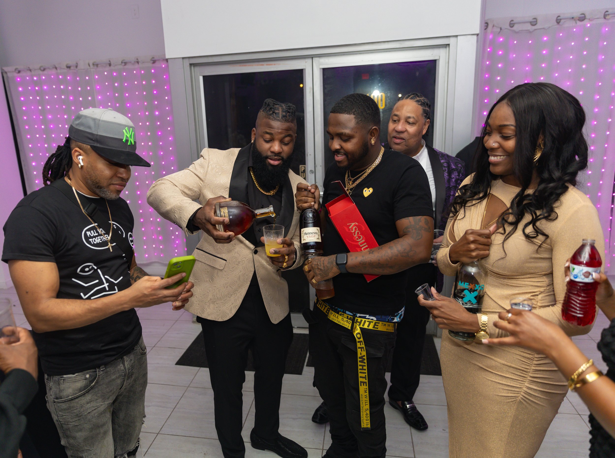 A group of five people gathered together at a party or celebration indoors. One person is pouring a drink into a glass, while others are looking at a phone or smiling. The background features purple LED lighting on the walls.