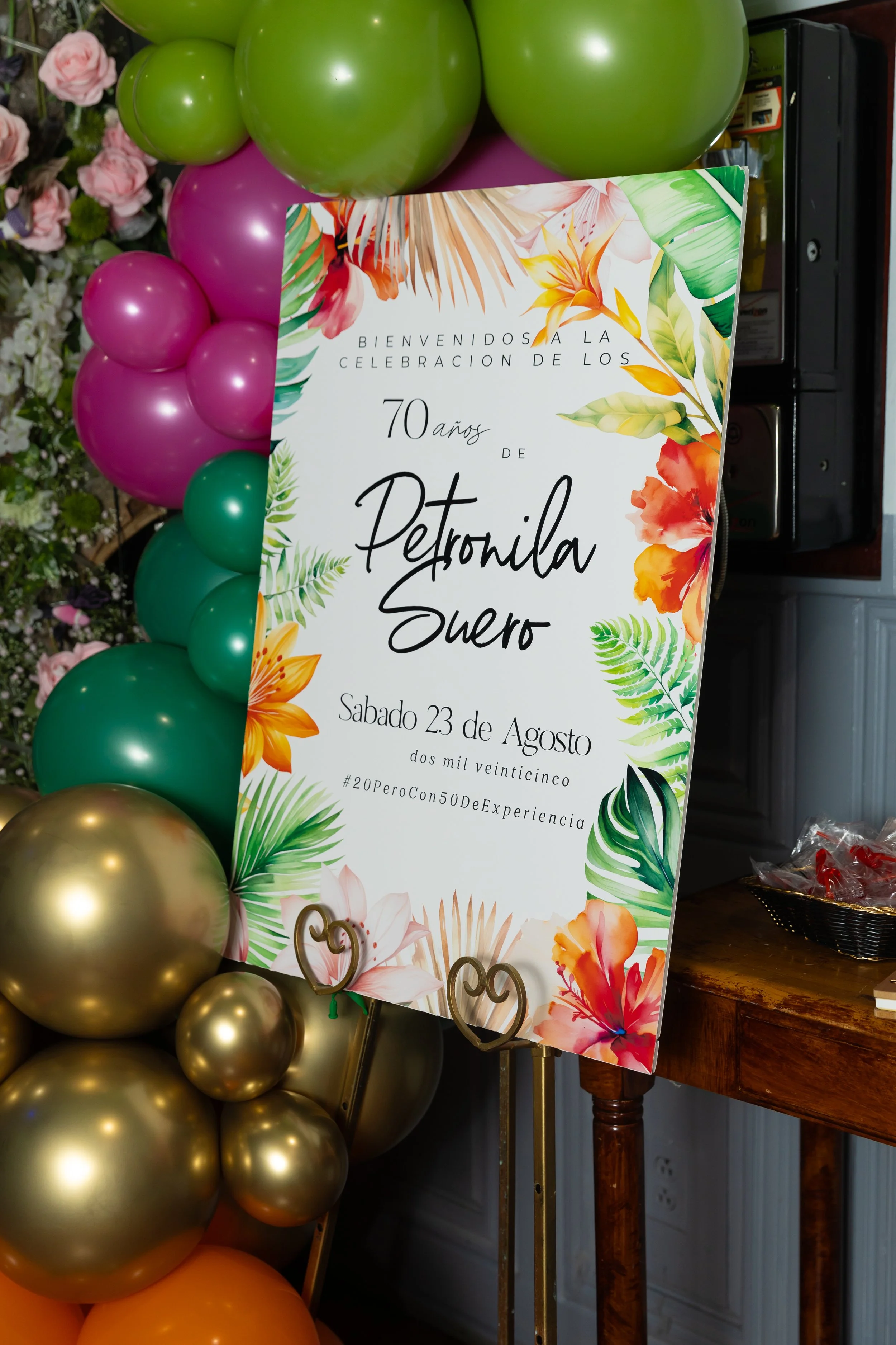 Celebration sign decorated with tropical flowers, surrounded by green, purple, gold, and orange balloons, for a 70th birthday party.