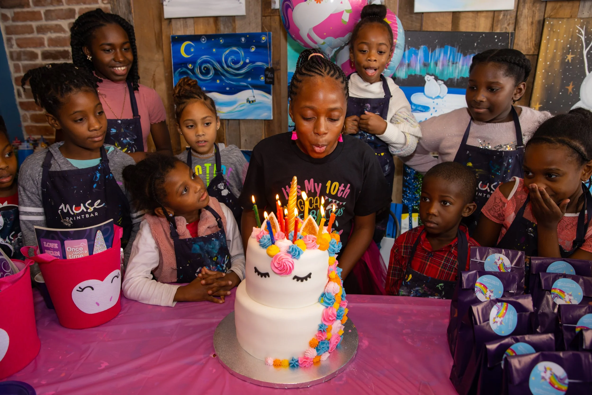 A birthday celebration with children gathered around a unicorn-themed birthday cake with lit candles. The children are wearing aprons and looking excited and curious, with some looking at the cake and others at the candles.