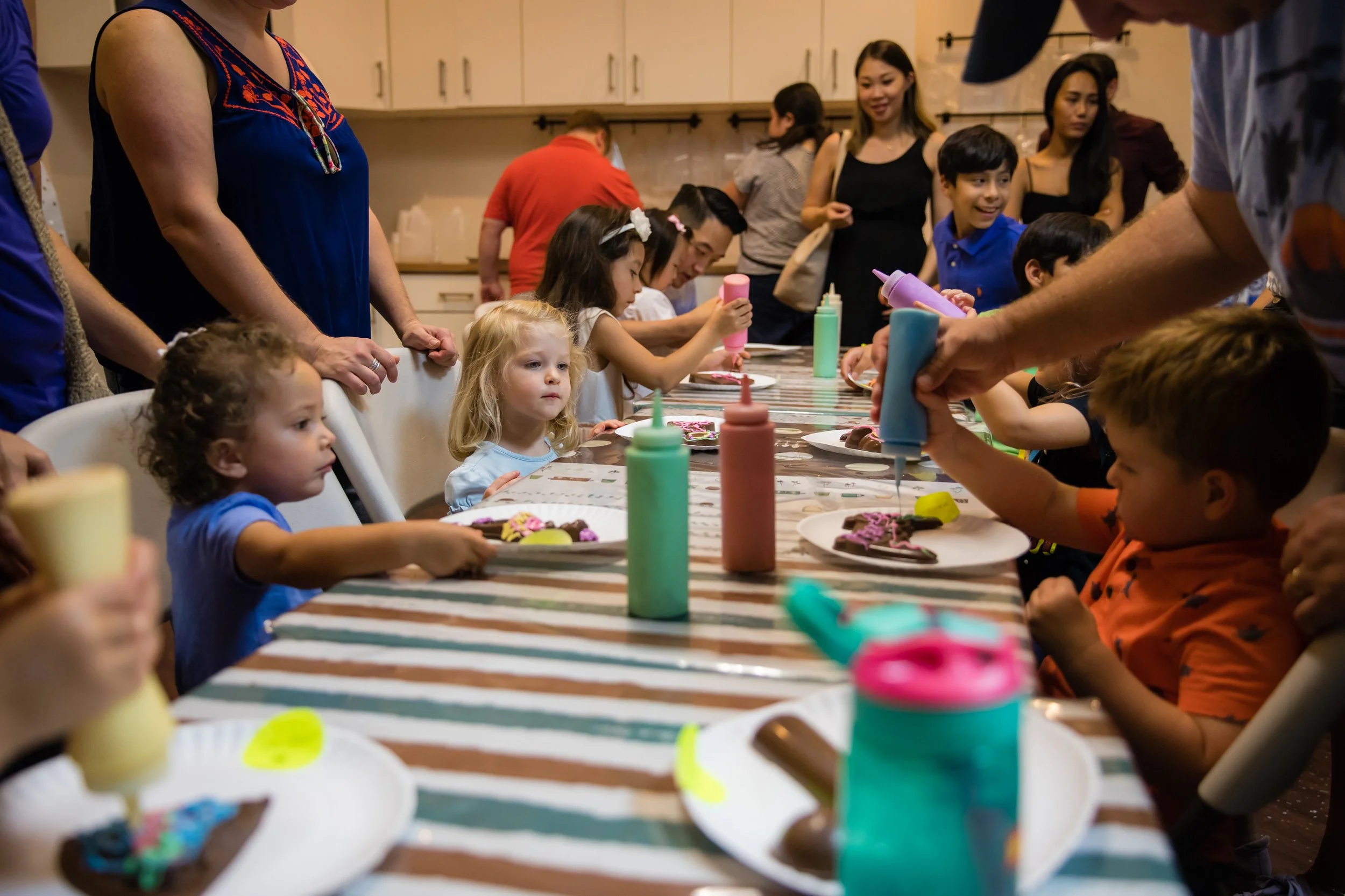People, including children and adults, gathered around a table at a party, decorating cookies with colorful icing and sprinkles.