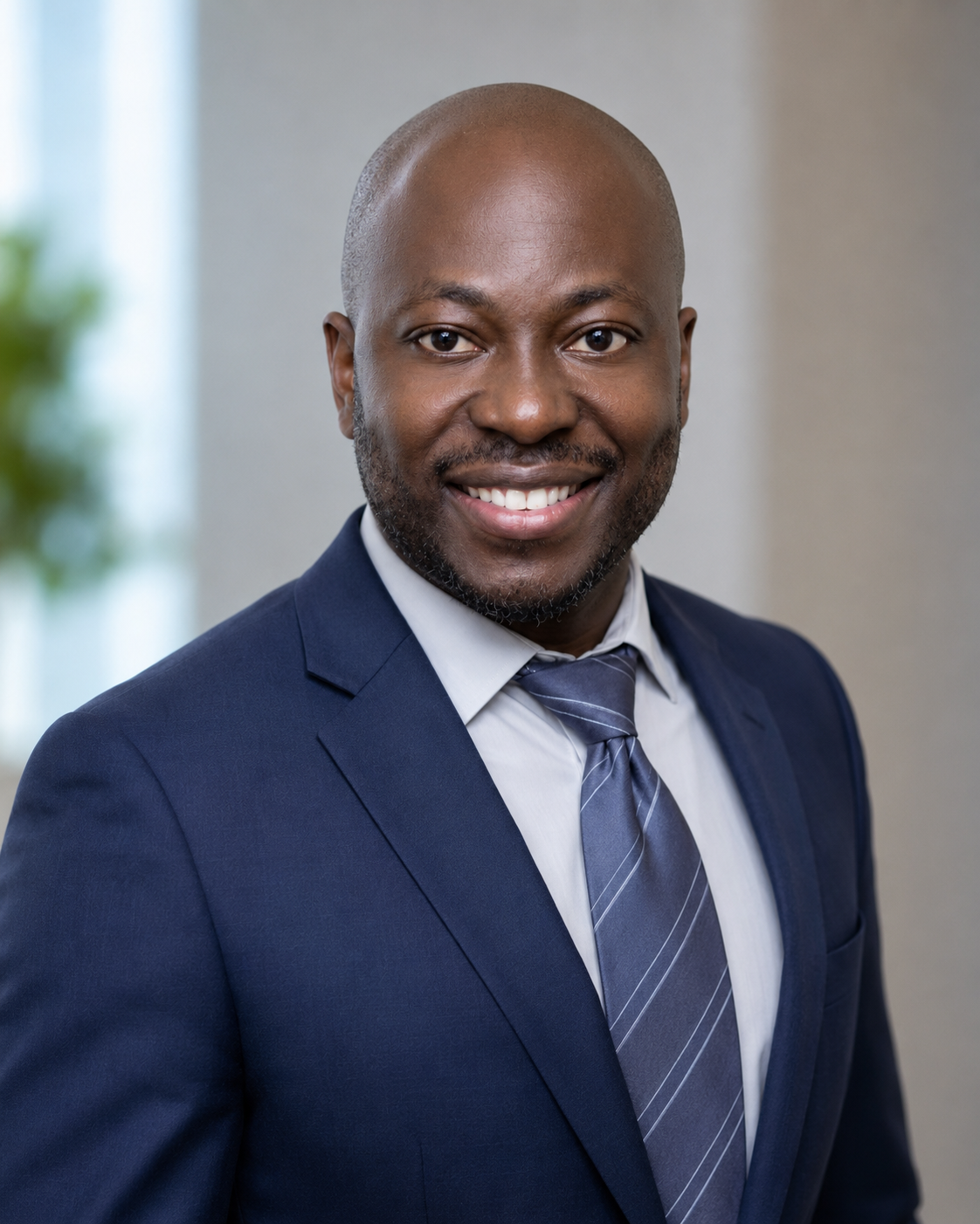 A smiling African American man in a navy blue suit, white shirt, and striped tie, standing indoors with a blurred background and a window.