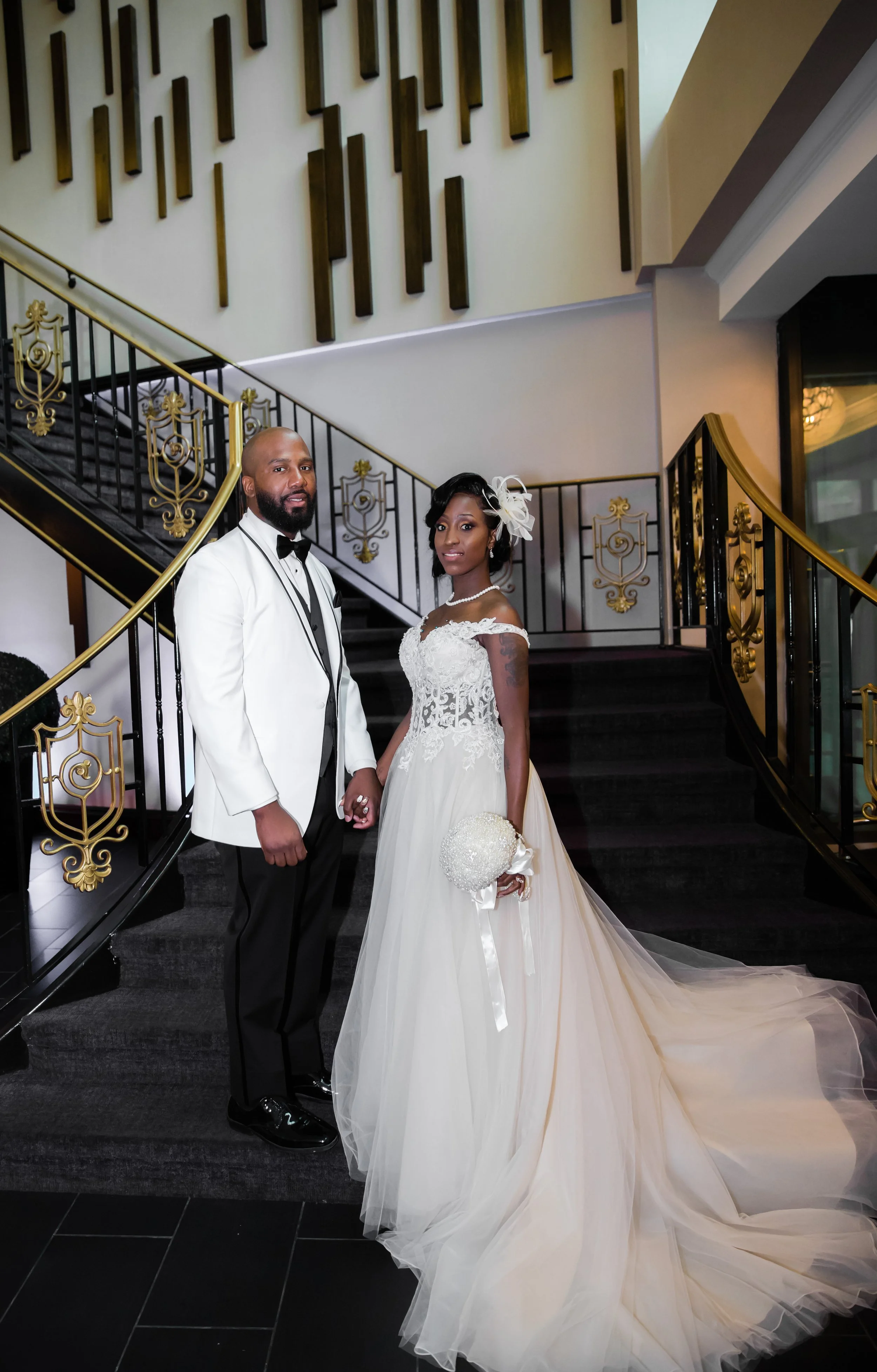 A bride and groom standing on a staircase inside a building. The bride is wearing a white wedding gown with lace details and a floral headpiece, holding a bouquet. The groom is wearing a white tuxedo with a black bow tie.