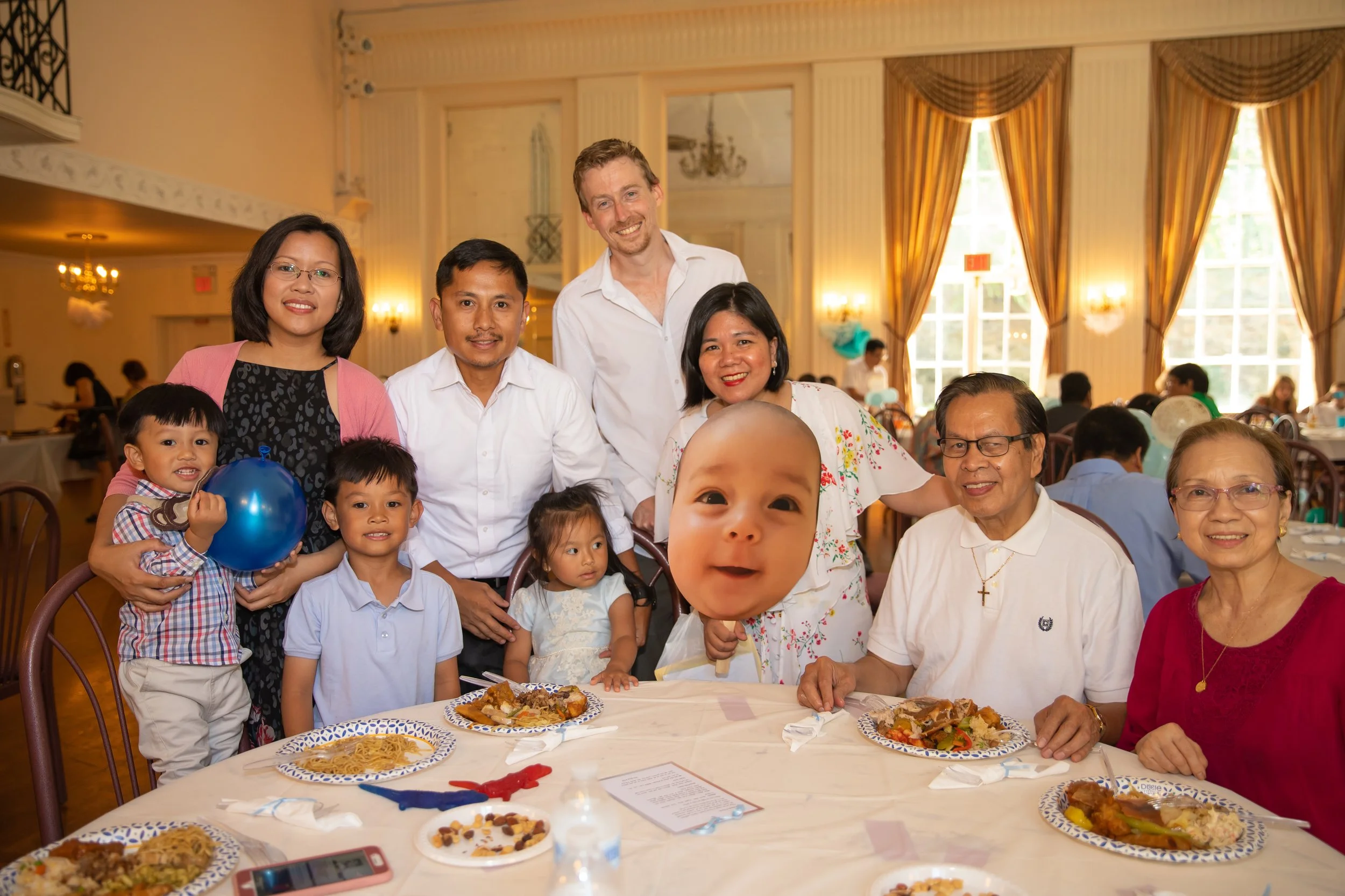 A diverse group of family and friends gathered around a table at a celebration, smiling at the camera, with food plates and decorations in a well-lit banquet hall.