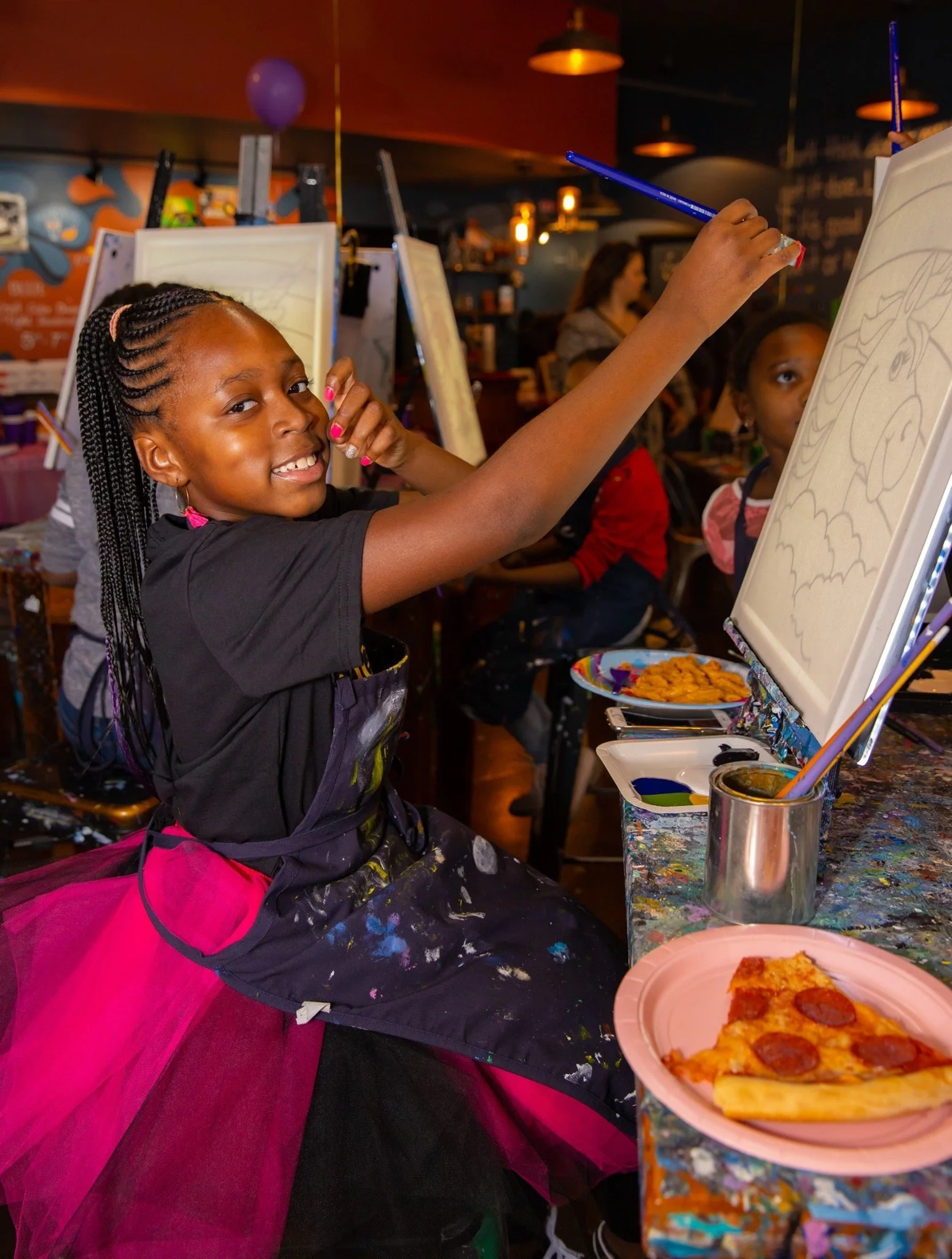 A young girl with braided hair smiling and drawing on a canvas at an art class, with other children in the background, in a cozy, decorated indoor space.