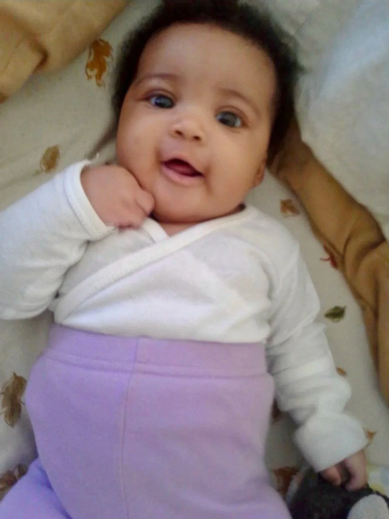 A baby with dark hair lying on a bed with a floral pattern, wearing a white long sleeve shirt and purple pants, looking up and smiling.