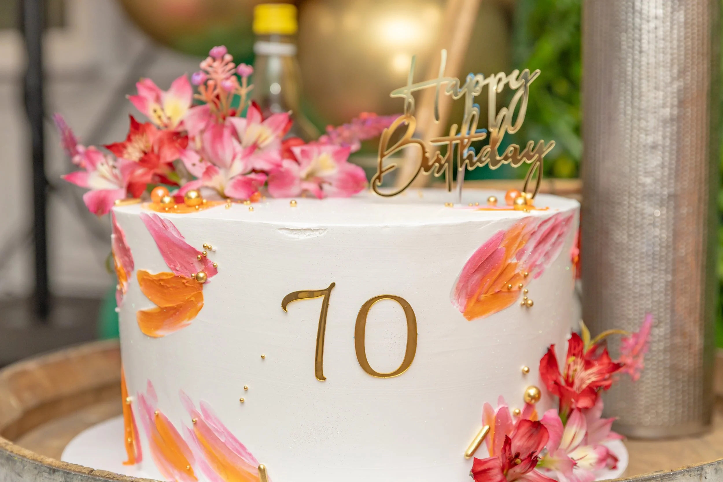 A decorated birthday cake with pink and orange flower accents, gold beads, and a gold 'Happy Birthday' topper. The cake has a smooth white frosting and a large gold number 10 on the front.