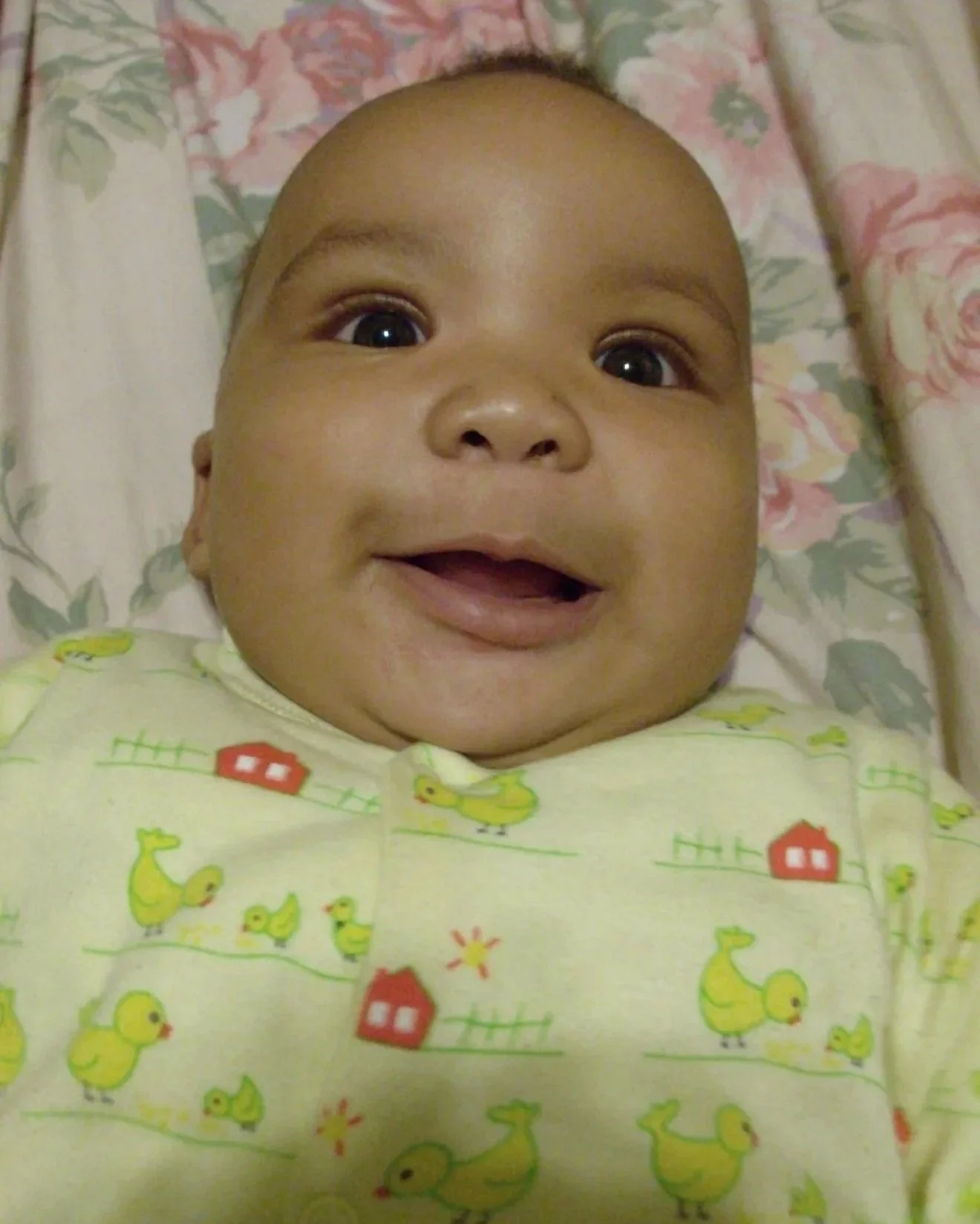 Close-up of a smiling baby with wide eyes lying in bed with a floral blanket in the background, wearing a yellow onesie with duck and house prints.