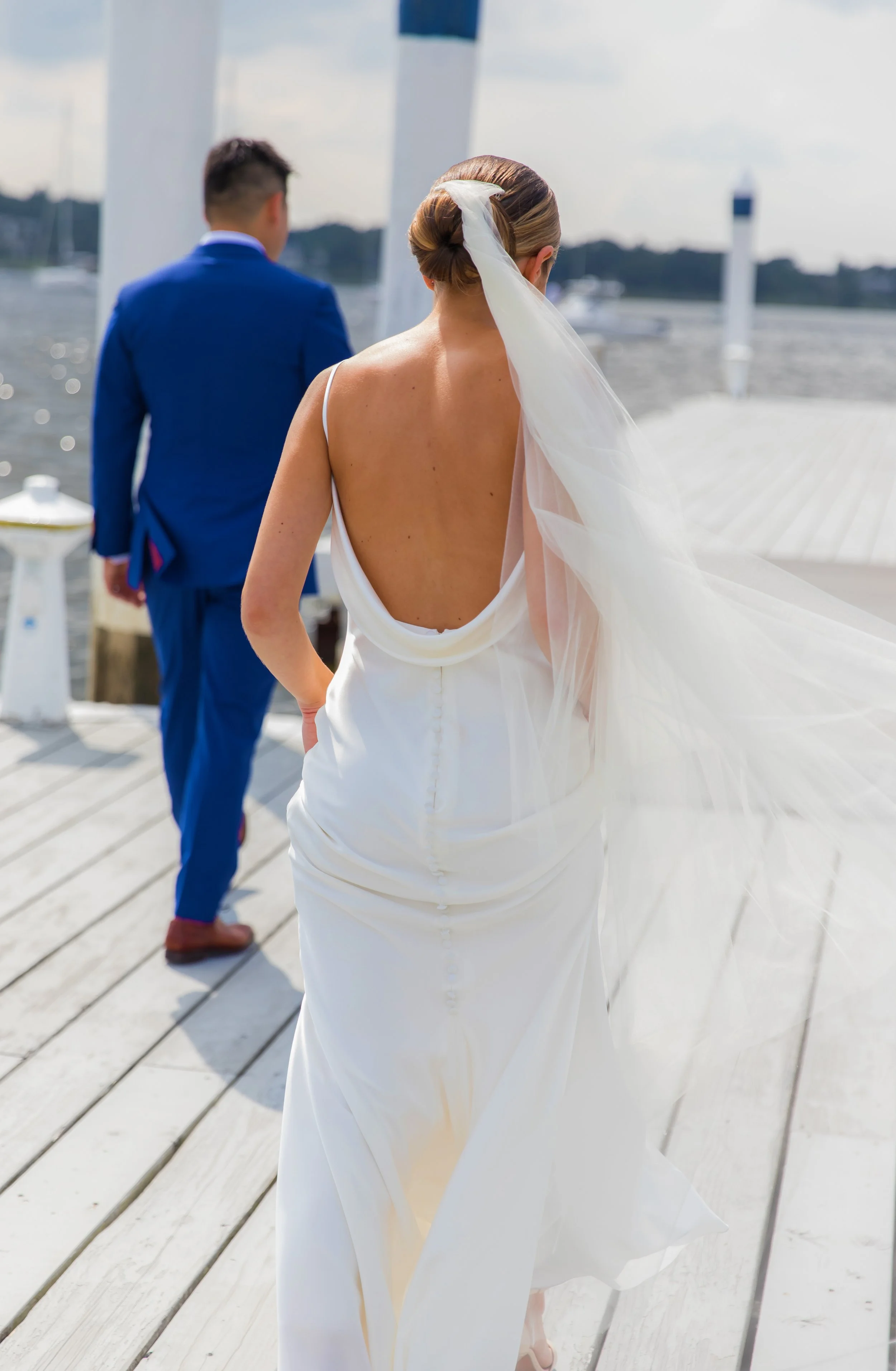 A bride with a backless white wedding dress and her hair styled in an elegant updo with a veil, standing on a wooden dock near the water, with a groom in a blue suit facing away in the background.