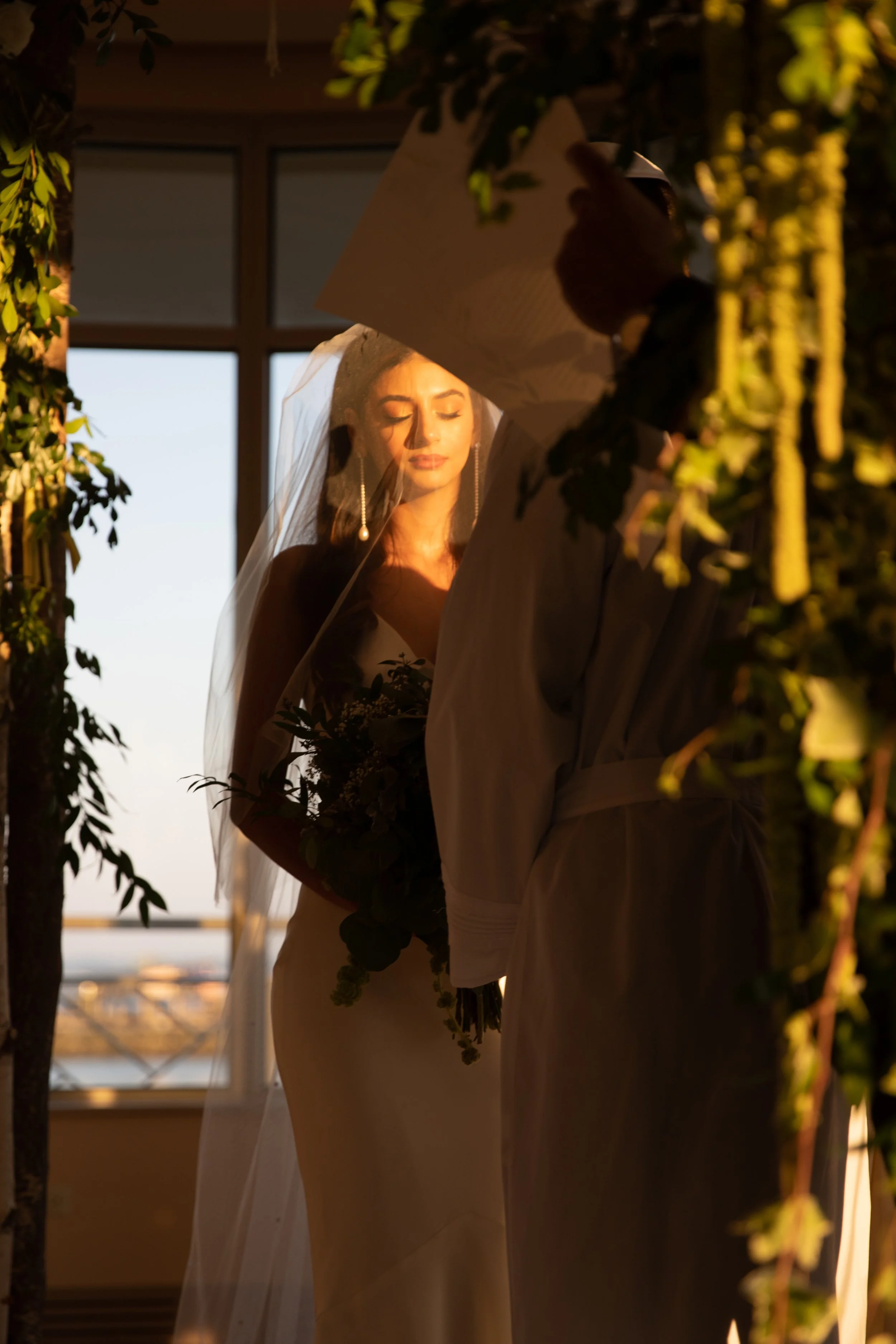 A bride standing with eyes closed, wearing a veil and earrings, holding a bouquet of flowers, surrounded by greenery, during sunset.