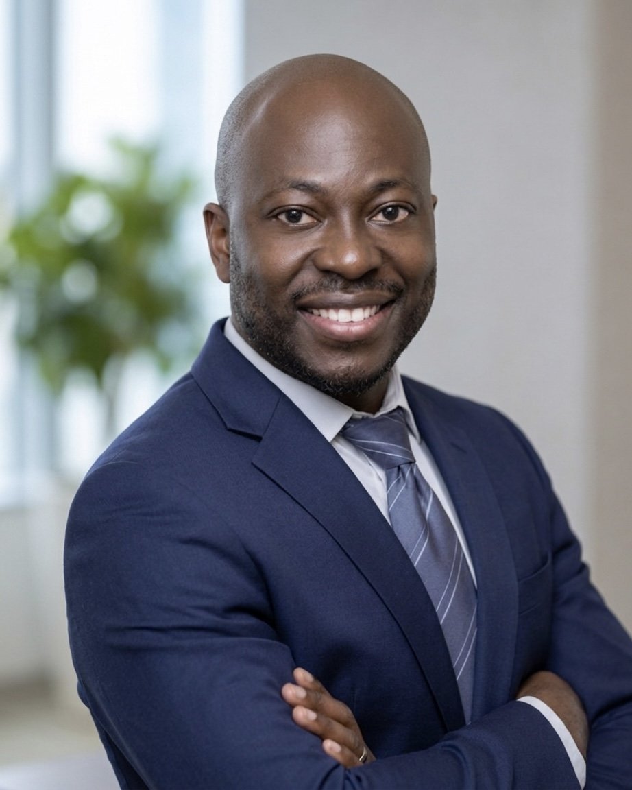 A professional African American man in a navy suit with a white shirt and striped gray tie, smiling with arms crossed in an office setting with a blurred background including a window and green plant.