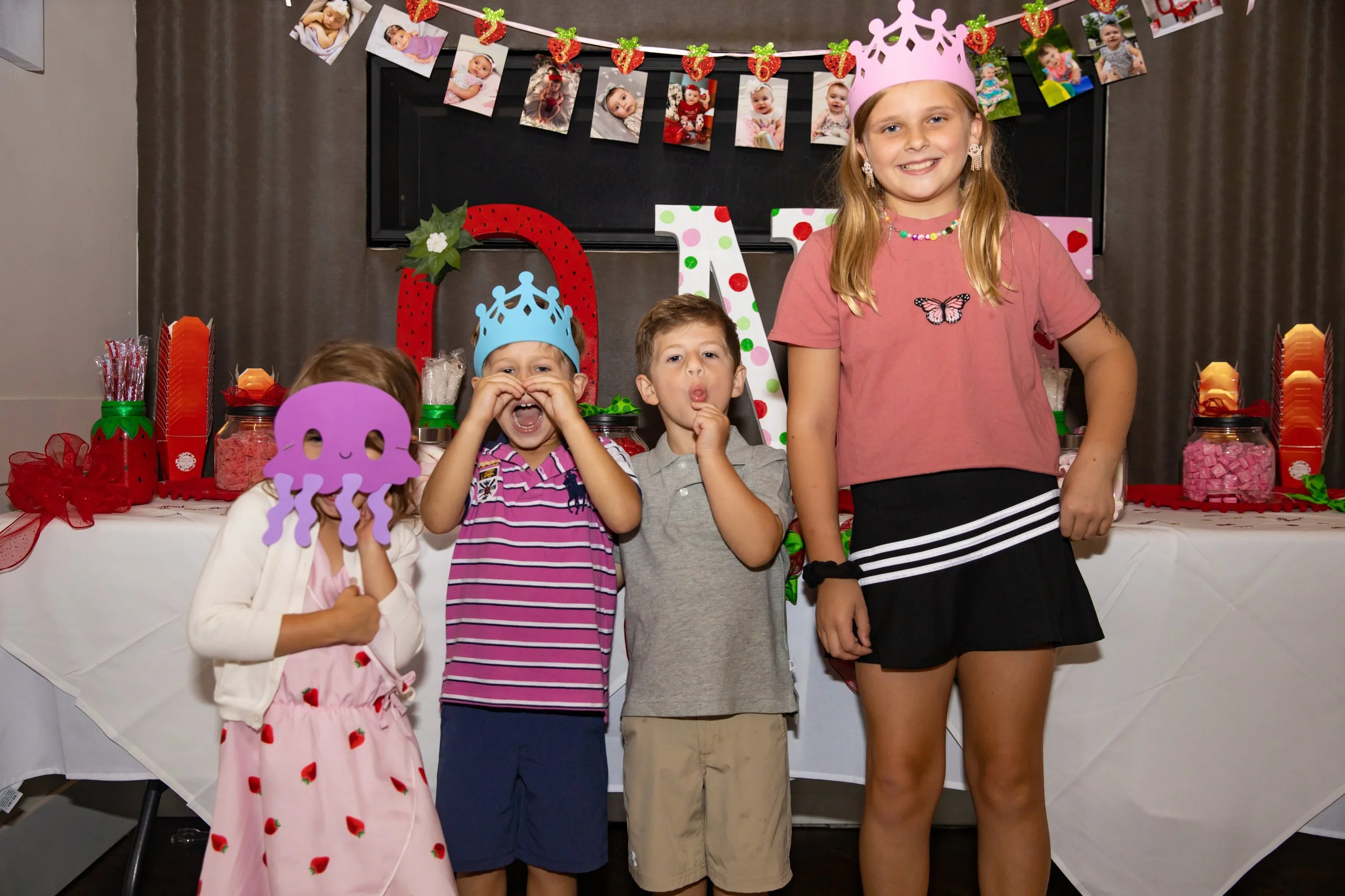 A girl with long blonde hair wearing a pink crown, smiling, standing next to three children at a birthday celebration. The children are wearing celebratory accessories, and the table is decorated with pink and red party items, candies, and a backdrop