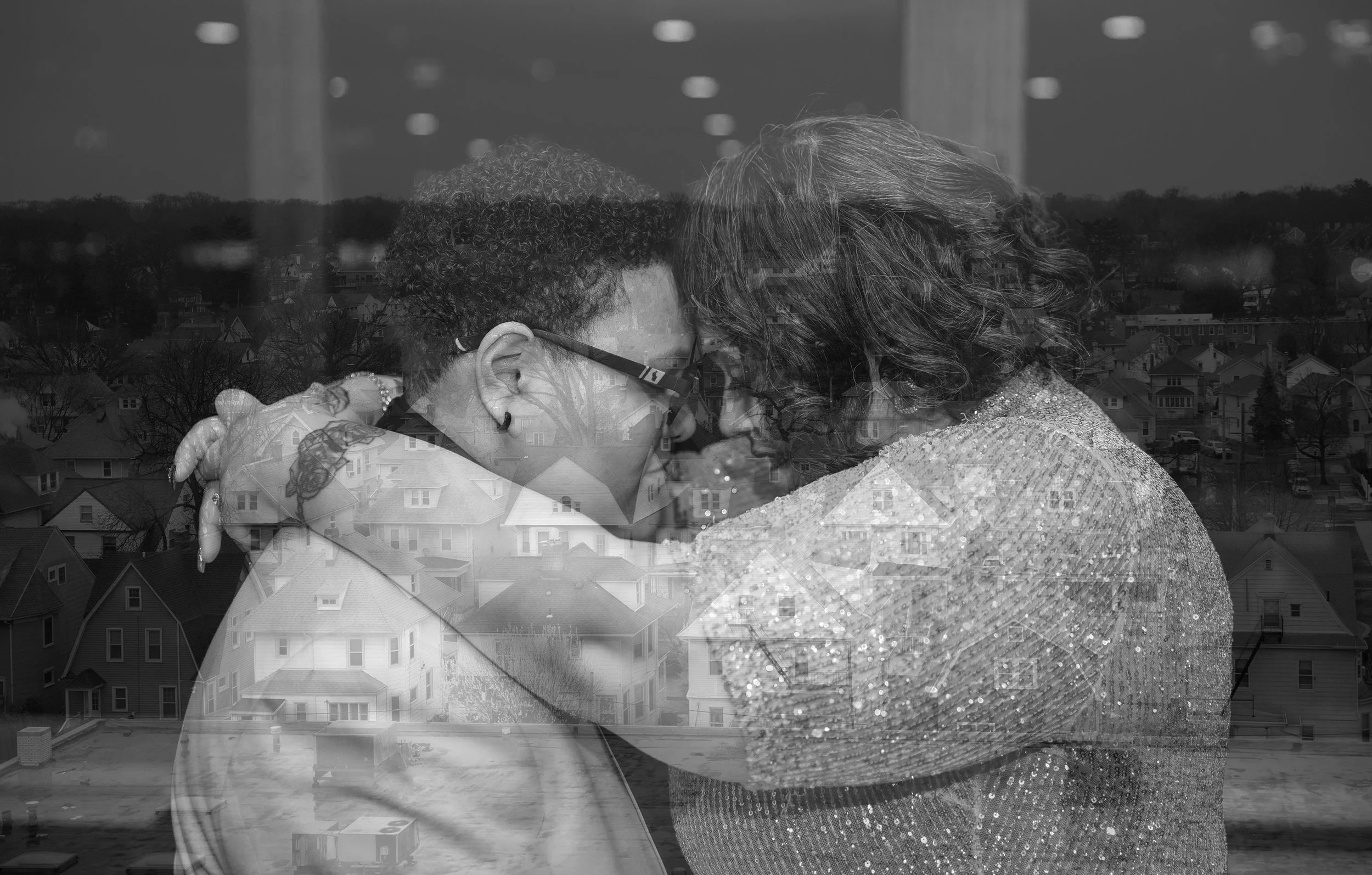 A black and white photo of a woman and a child sharing a tender moment, with their foreheads touching and eyes closed. The woman has curly hair and is wearing a sparkly top, while the child has short, curly hair and wears glasses. The photo has a dou